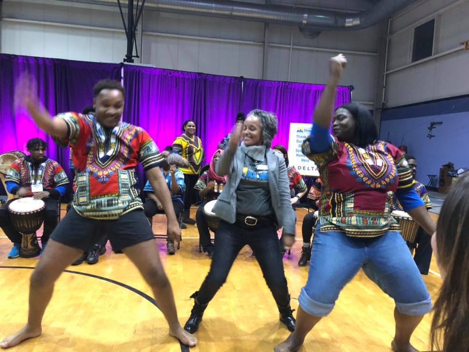 Three women dancing with joyful expressions in a gymnasium, colorful clothing, purple curtain background, and other people in the background.