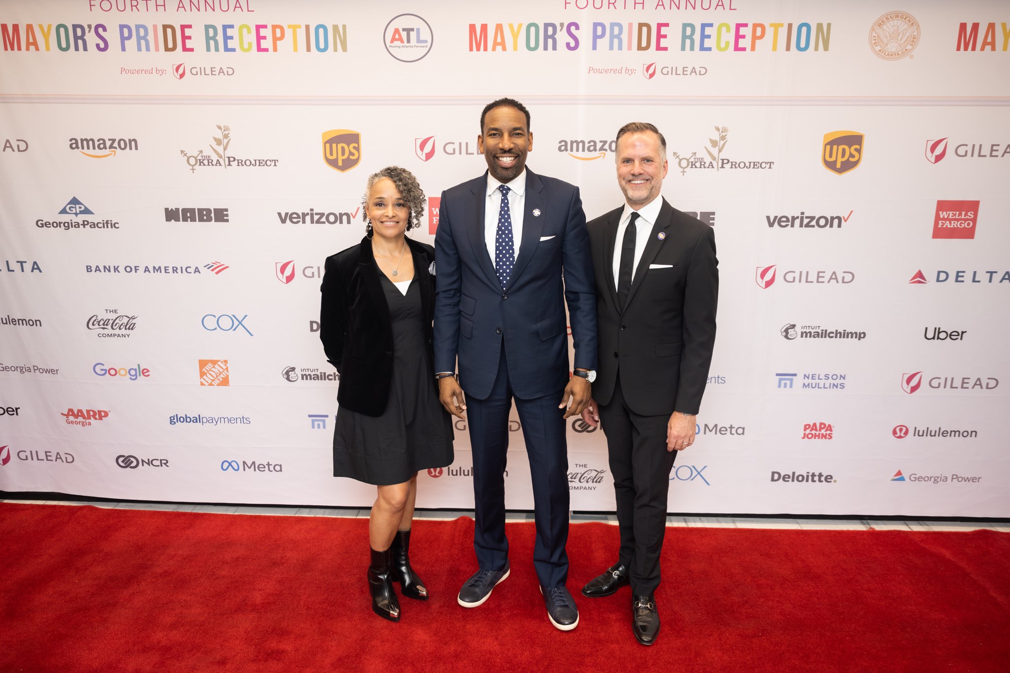 Three people standing on a red carpet in front of a backdrop with logos, smiling and holding hands.