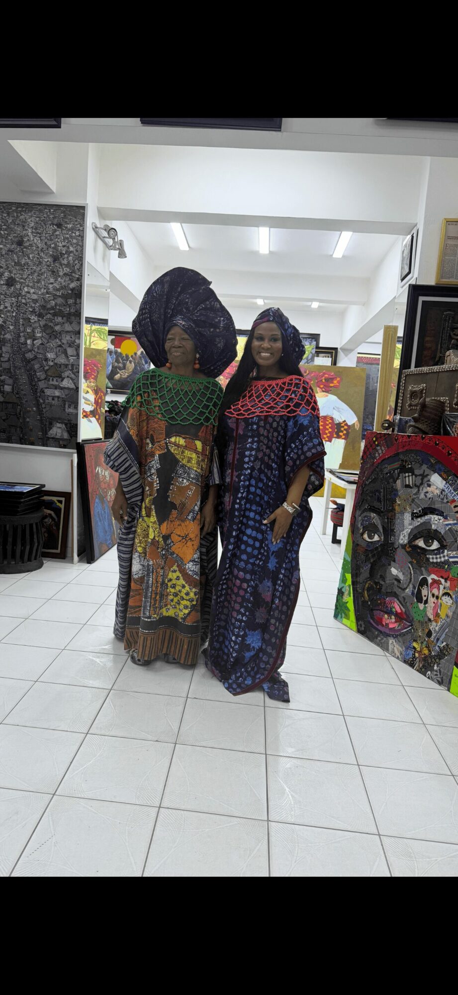 Two women in colorful traditional dresses stand indoors, smiling, with artwork displayed around them.