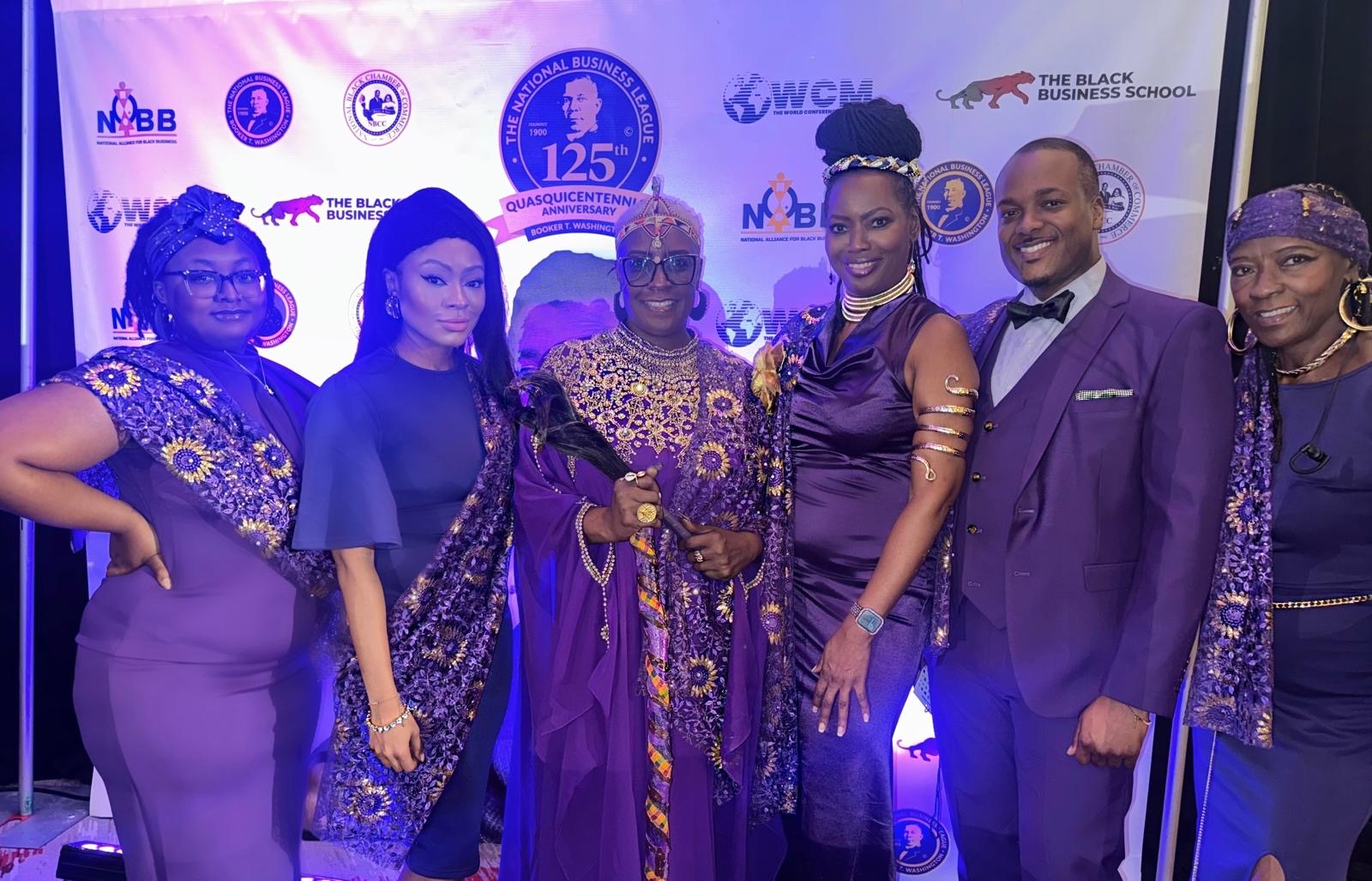 Group of seven people dressed in purple and blue attire, standing in front of a backdrop with logos and text.