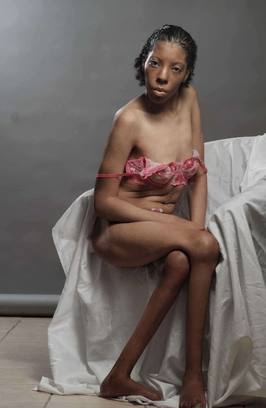 Young woman sitting on a white cloth-covered surface, wearing pink lingerie, with a neutral gray background.