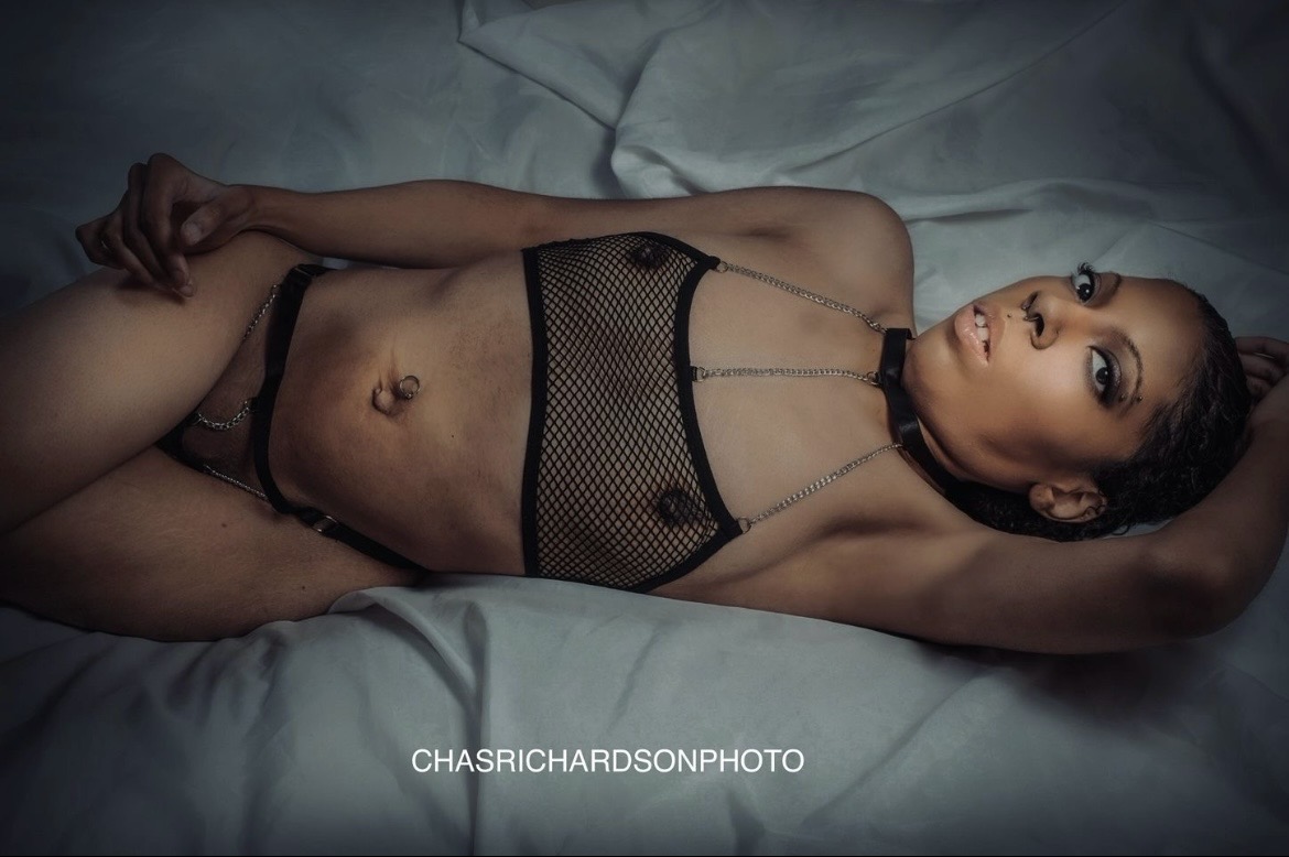 Woman lying on bed wearing black mesh lingerie, with arm behind head, looking at camera, dark background, soft lighting.