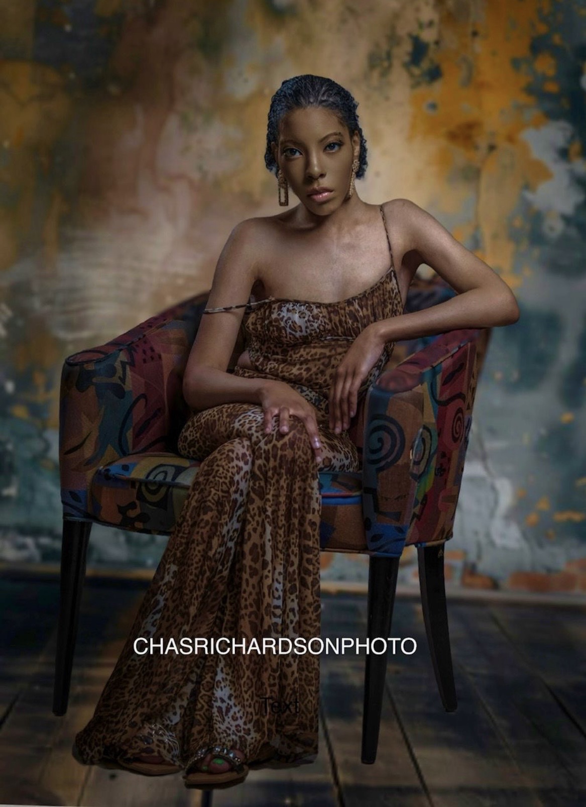 Young woman in leopard print dress sitting on a colorful patterned chair against a textured background.