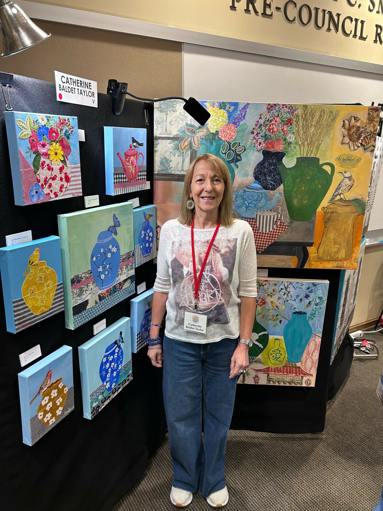 Woman standing next to colorful artwork display at an event, with paintings of flowers, animals, and abstract designs.