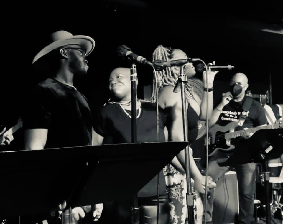 Group of five musicians performing on stage with microphones and instruments, in black and white.