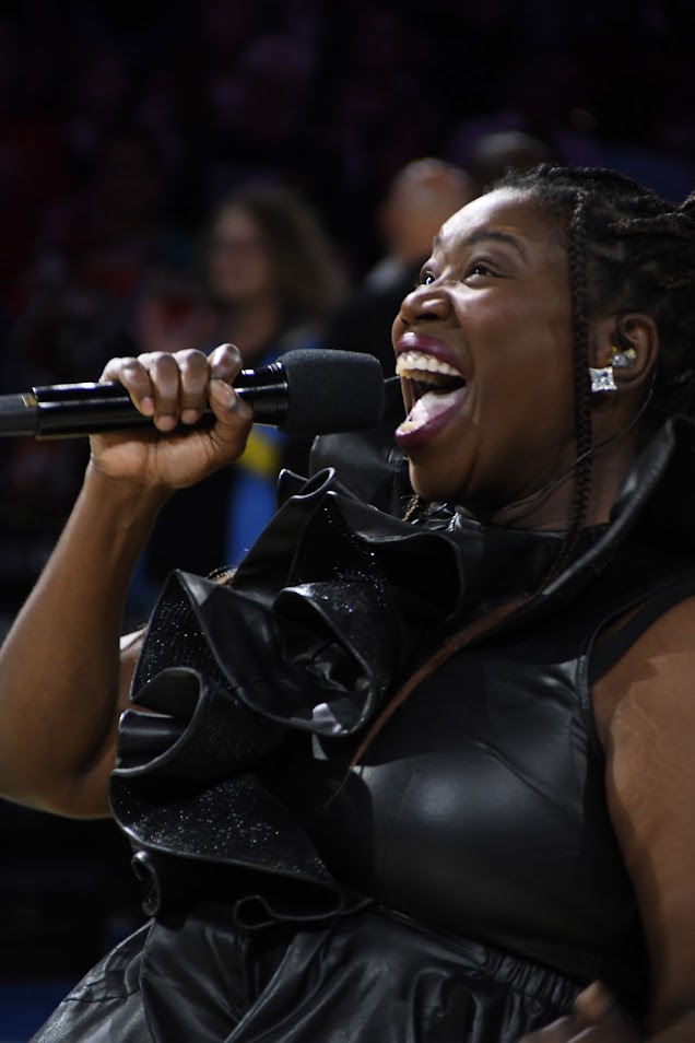 Woman singing into a microphone, smiling, with an audience in the background.