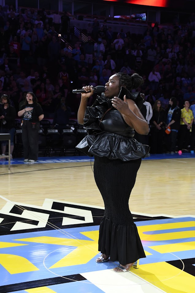 A woman singing into a microphone on a basketball court with a crowd in the background.