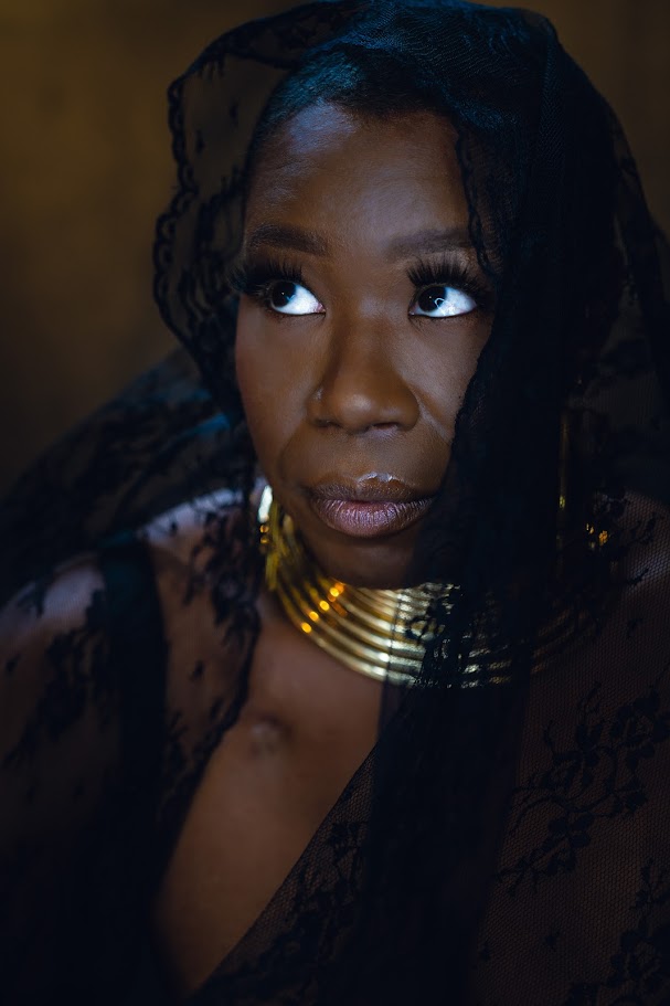 Close-up portrait of a woman with dark skin, wearing a black lace head covering and gold jewelry, looking to the side.