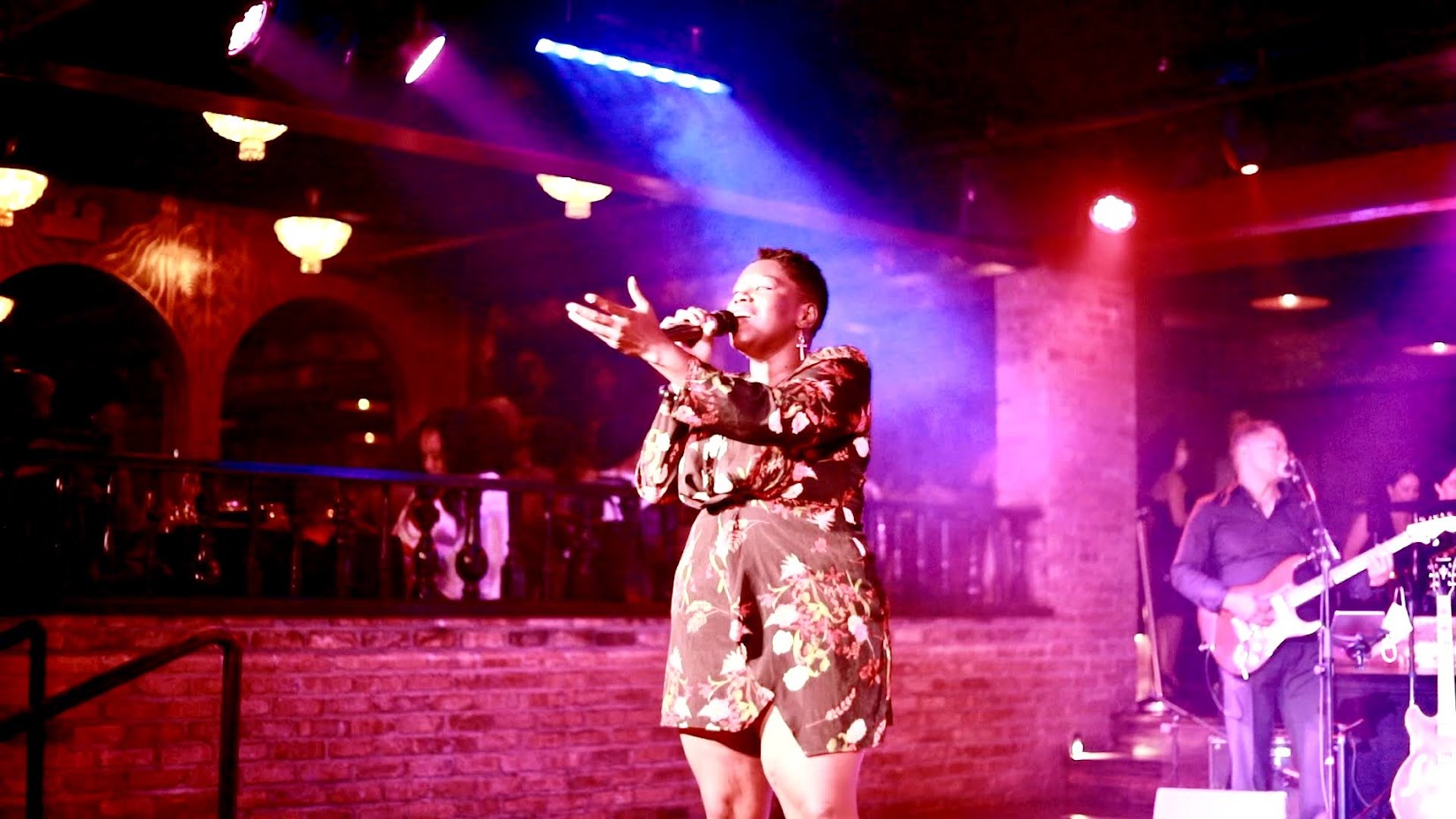 Performer singing on stage with colorful lights, a guitarist on the right, and audience in the background.