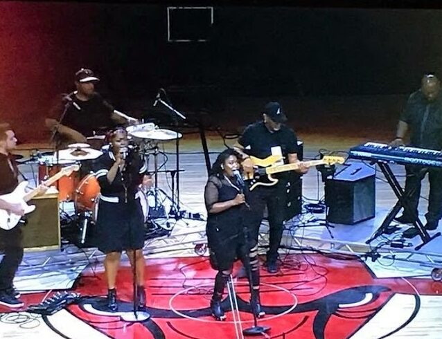 Band performing on stage with drums, guitars, keyboard, and microphones, on a basketball court with a red logo.
