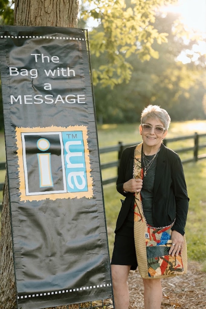 Woman standing outdoors holding a colorful bag, smiling, with a sign that reads 'The Bag with a MESSAGE' and a logo, trees, and sunlight in background.