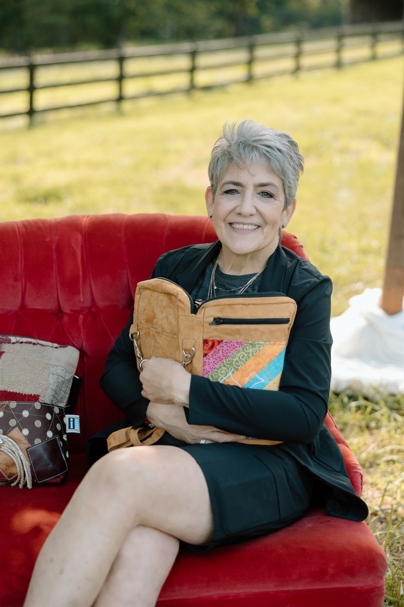 Older woman with short gray hair smiling, sitting on a red outdoor sofa, holding a colorful book, in a grassy area.