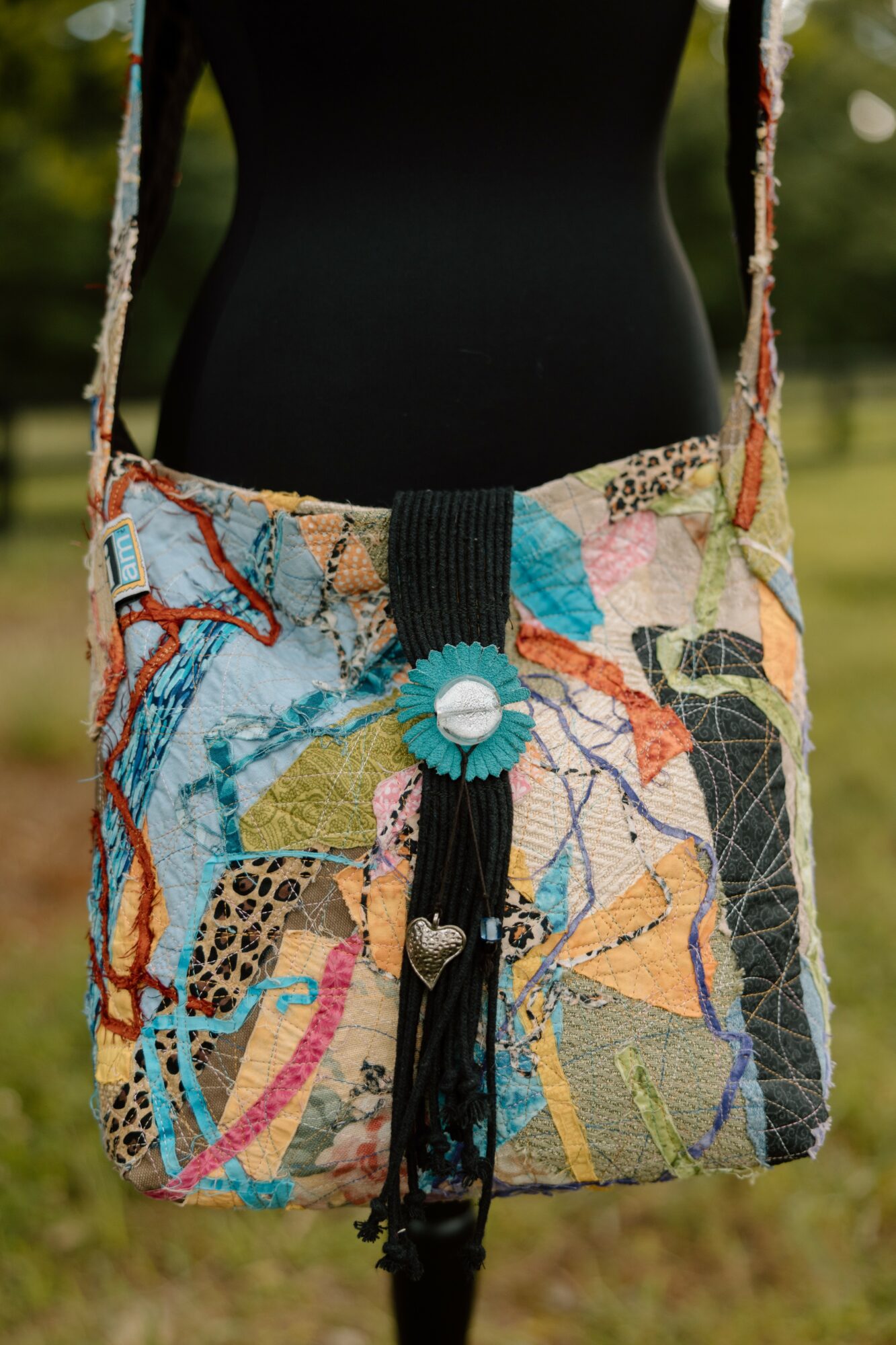 Person wearing a colorful, embroidered shoulder bag with a black tassel, outdoors with blurred greenery in background.