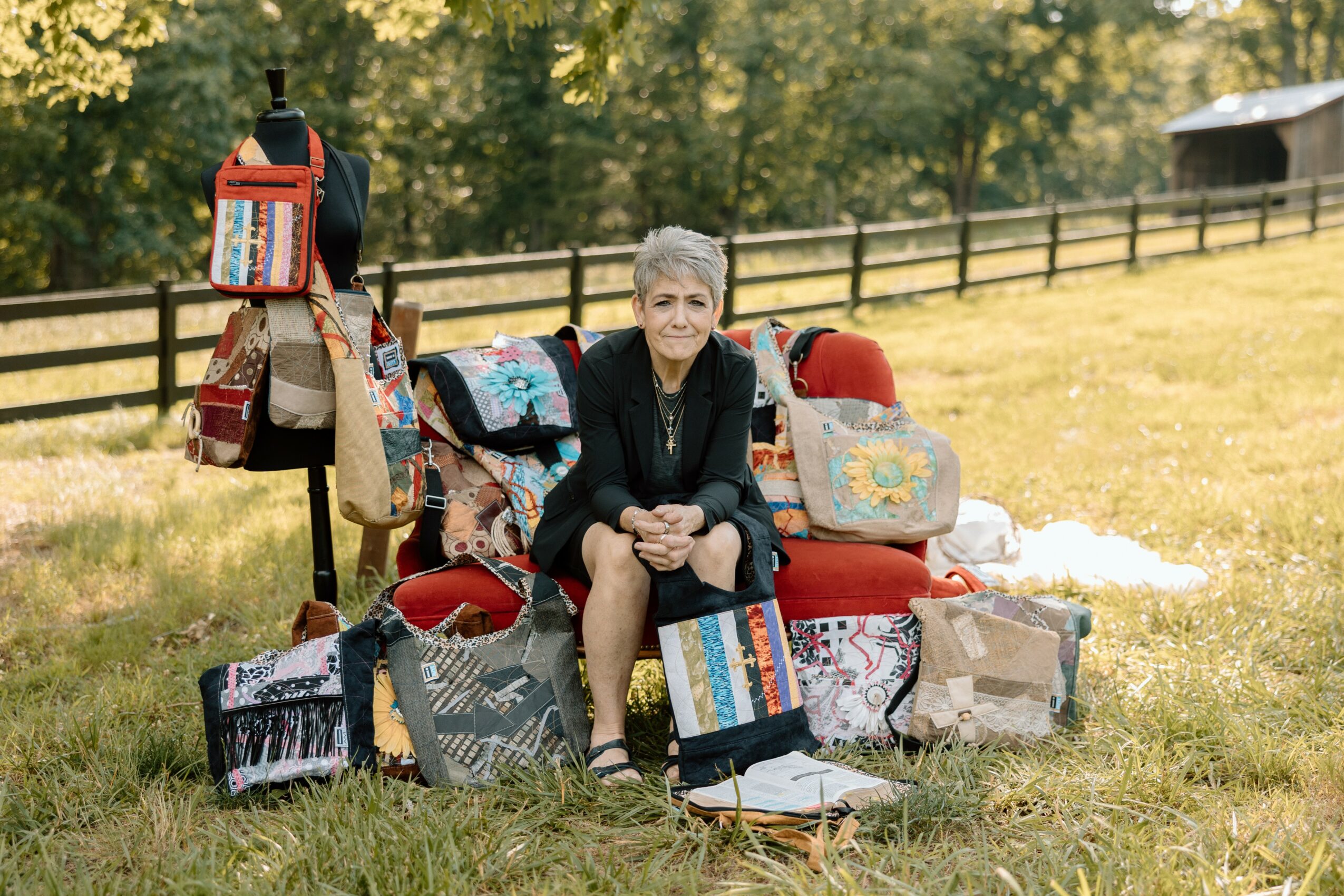 Older woman sitting on a red bench surrounded by bags and quilts outdoors in a grassy field.