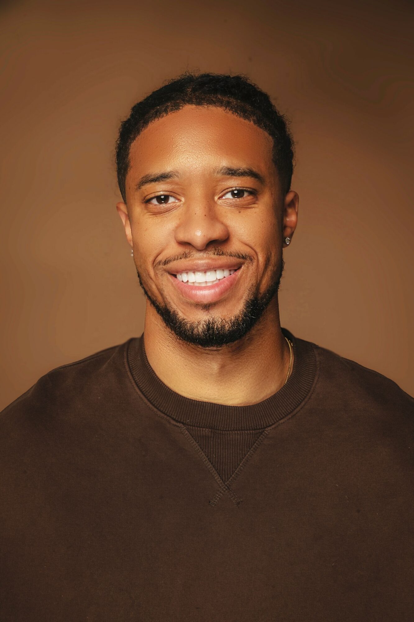 Portrait of a smiling man with short curly hair and a beard, wearing a dark shirt, against a brown background.