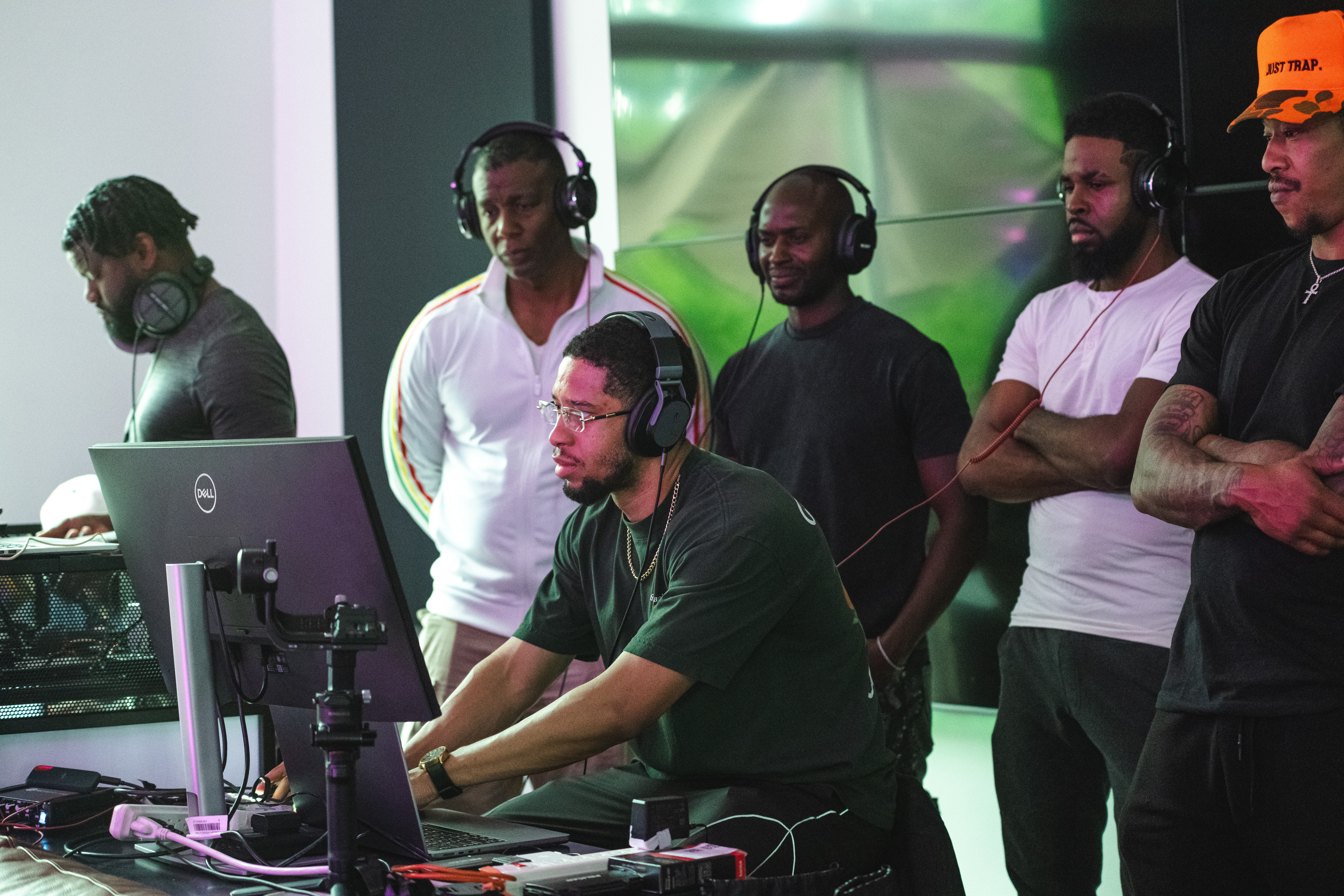 Group of five men, one using a computer with headphones, others watching, in a modern room with green lighting, casual clothing.
