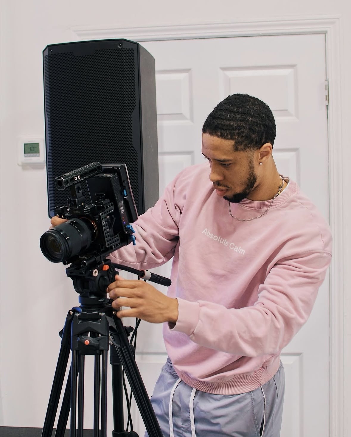 Young man operating a professional camera on a tripod indoors, wearing a pink sweatshirt and gray pants.