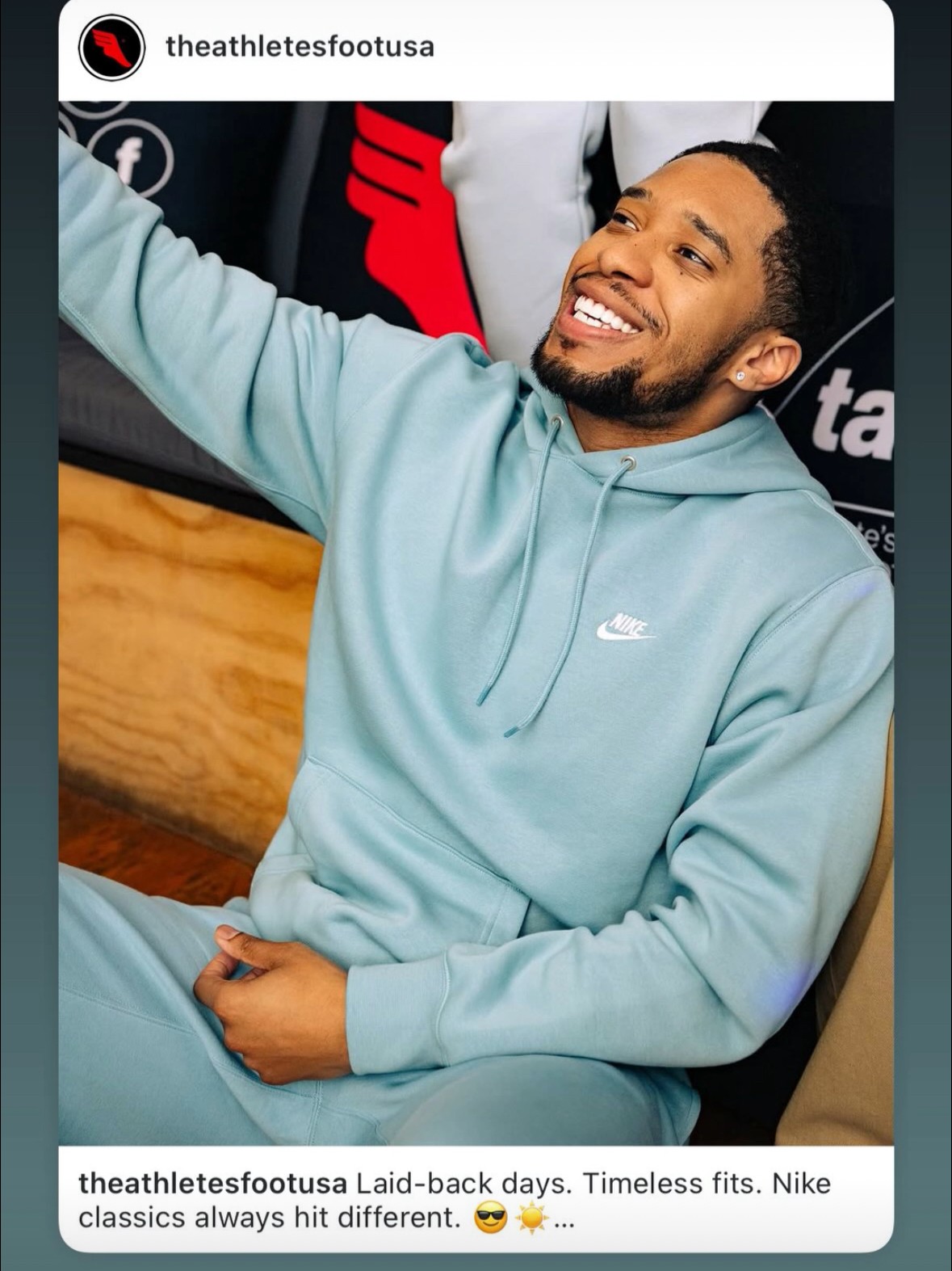 Smiling man in light blue Nike hoodie sitting and taking a selfie, background includes a wooden surface and sports-related posters.