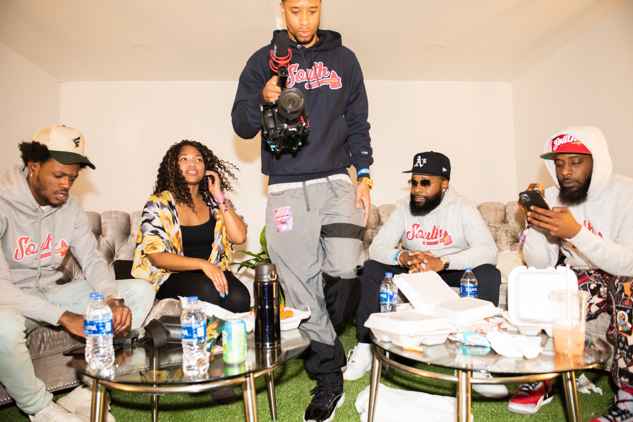 Five people sitting around a table with drinks and snacks, one person standing in the center holding a camera, in a room with white walls.