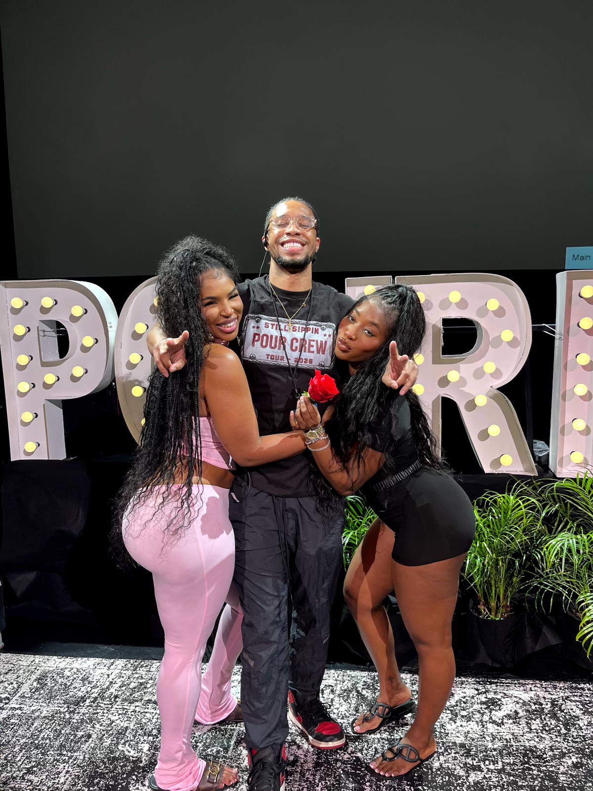 Three people smiling and hugging in front of large decorated letters, with plants nearby, on a stage.