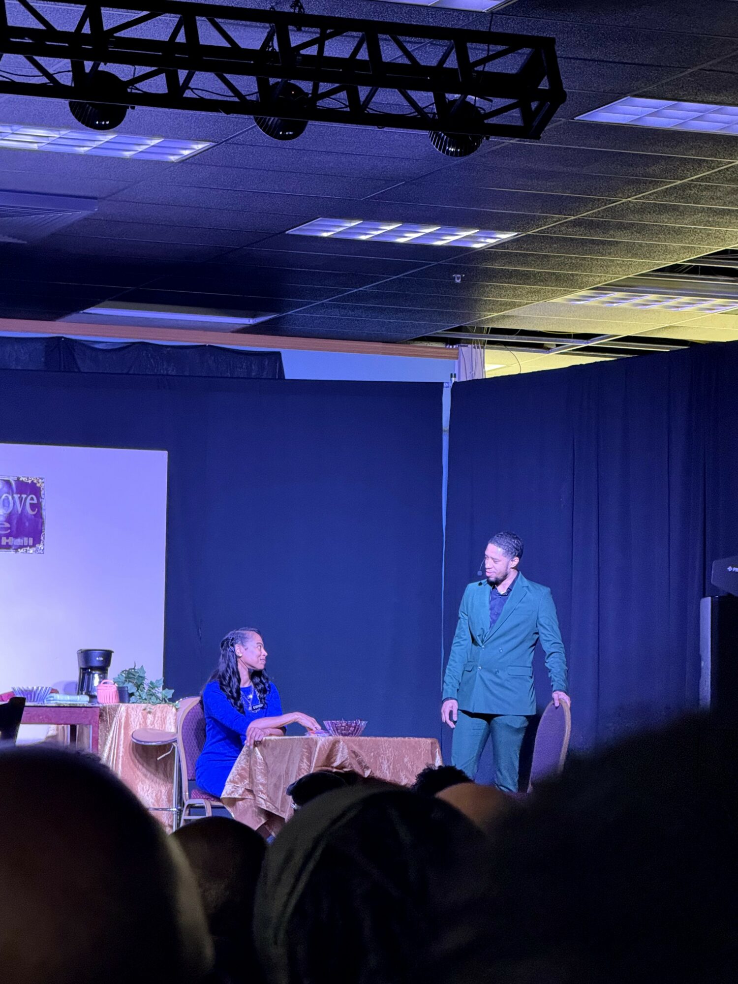 Two women on stage, one standing and one sitting at a table, with a dark curtain background and stage lighting.