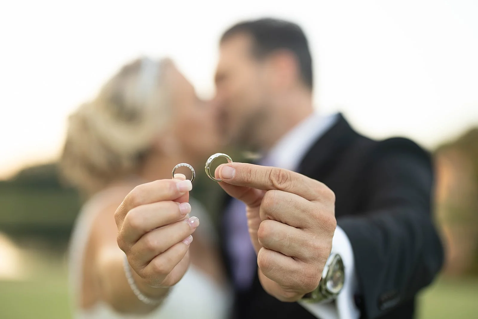 Two people holding wedding rings close to the camera, with a blurred couple kissing in the background outdoors.