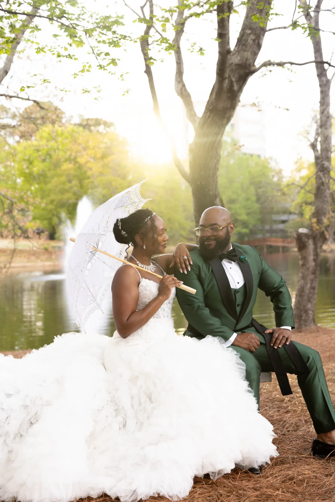 Bride and groom sit outdoors by a lake, with sunlight filtering through trees, bride holding a parasol, both smiling.