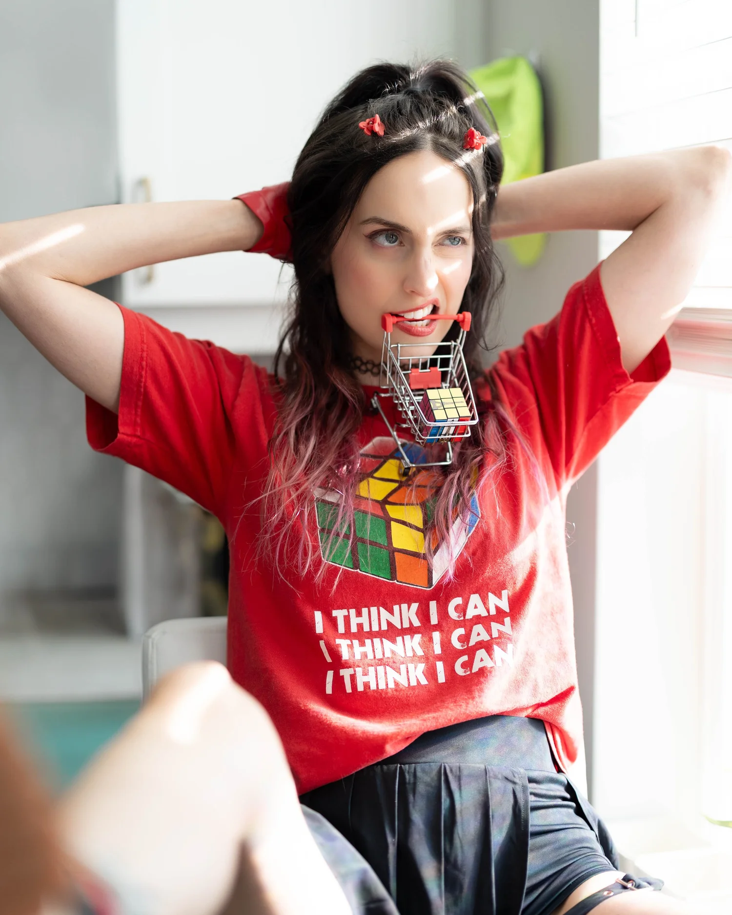 Woman with dark hair and red clips, wearing a red T-shirt and black skirt, holding a shopping cart in her mouth, smiling.