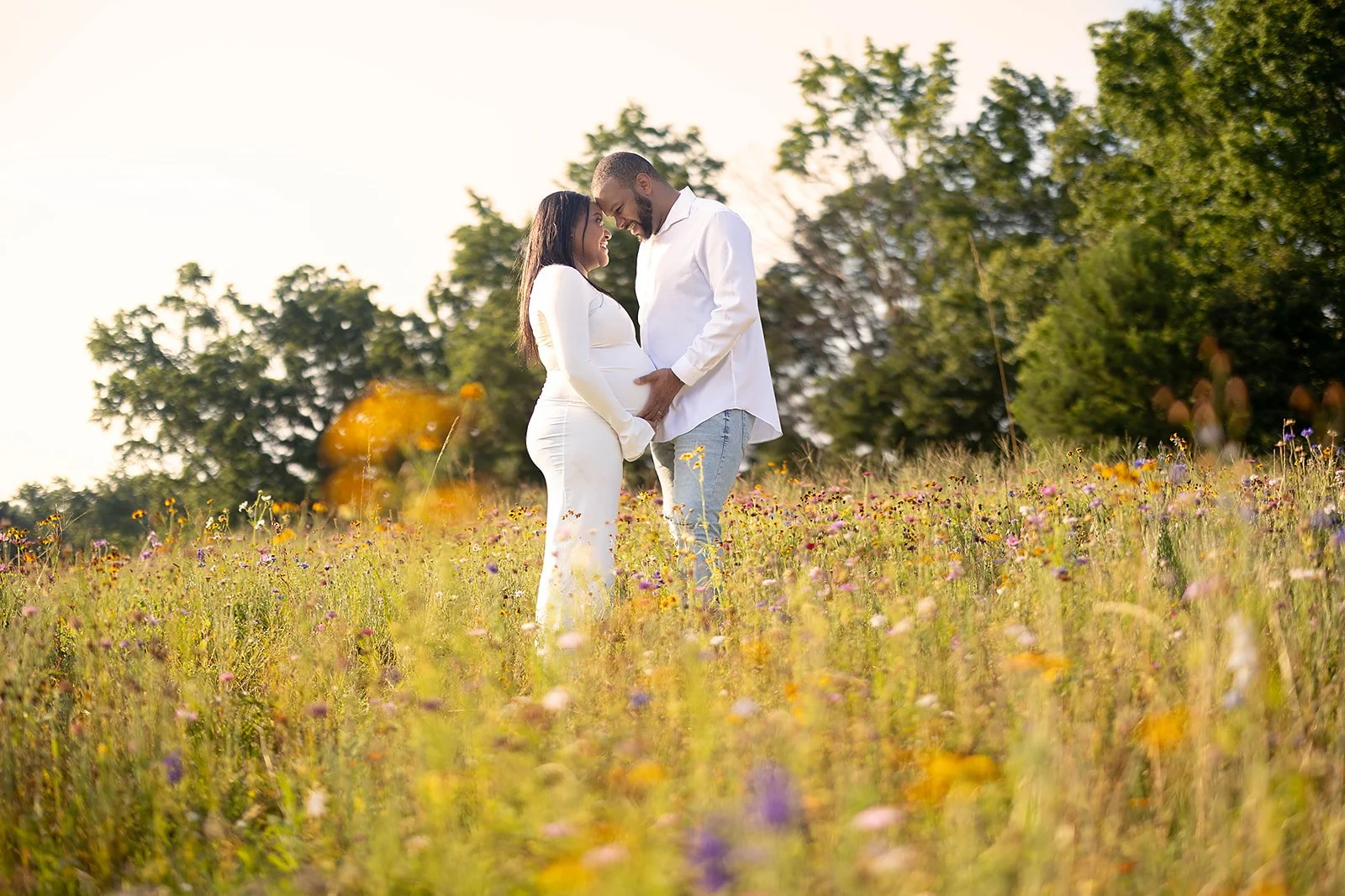 A couple stands close together in a field of wildflowers, holding hands and touching foreheads outdoors during daytime.