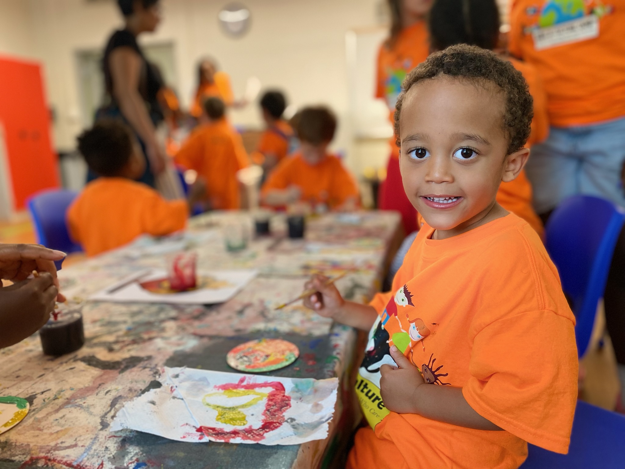 Young child with curly hair smiling, wearing an orange shirt, at a table with colorful artwork, in a lively indoor setting.