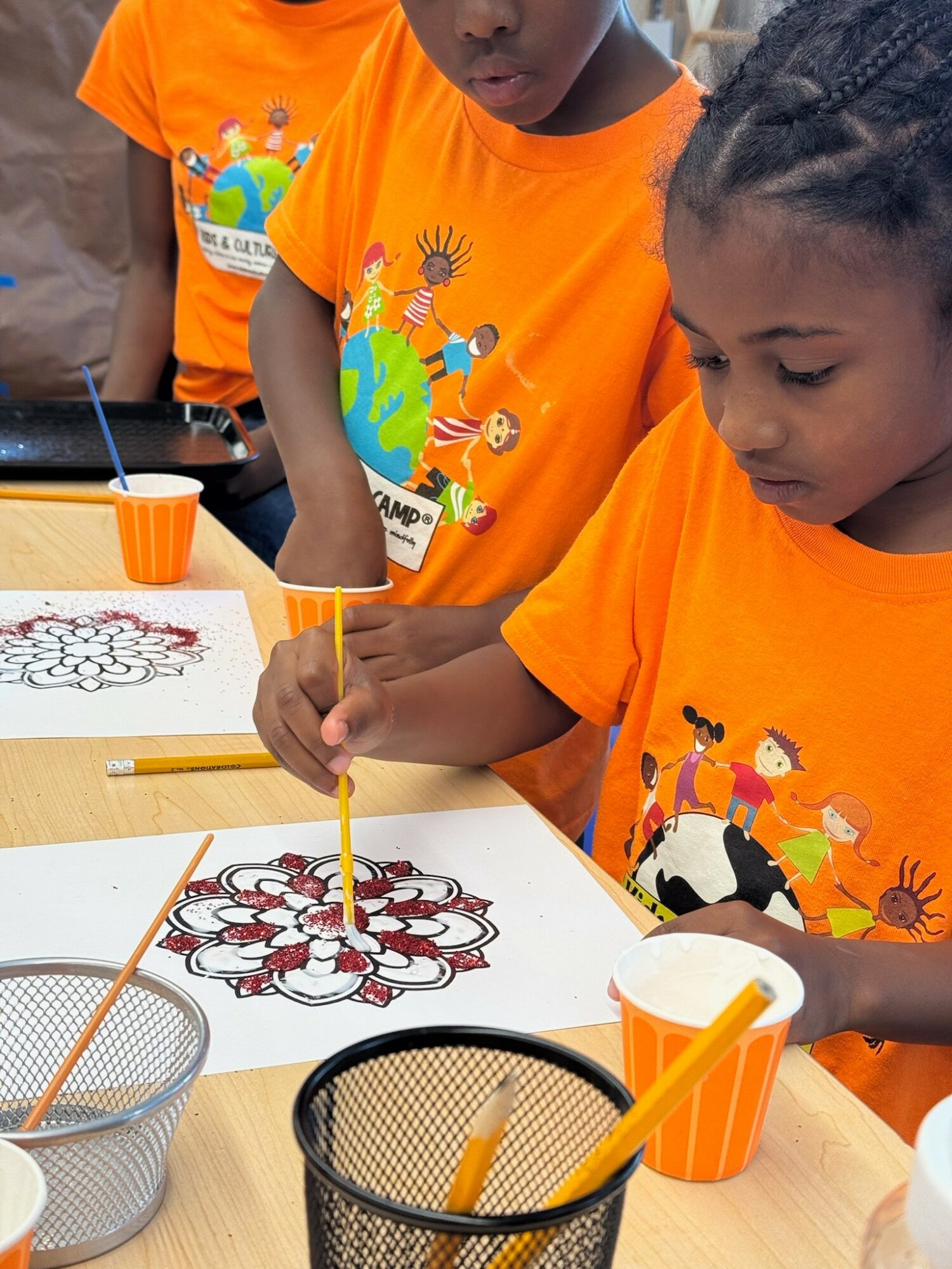 Children in orange shirts drawing and coloring on paper at a table, with art supplies nearby.