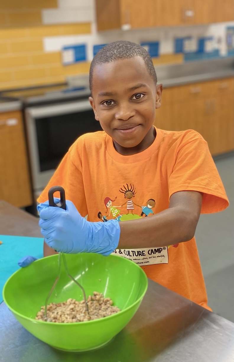 Young boy with short hair wearing an orange T-shirt and blue glove, stirring a bowl of food in a kitchen.