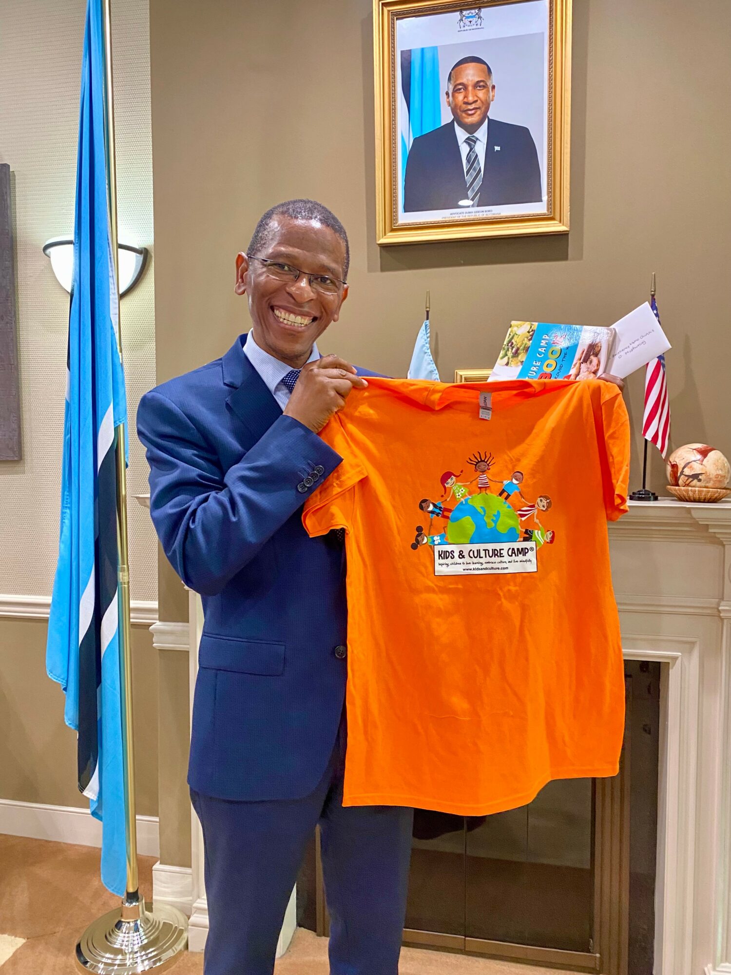 Smiling man in a suit holding an orange T-shirt with a colorful logo, standing in a room with a framed photo and flags.