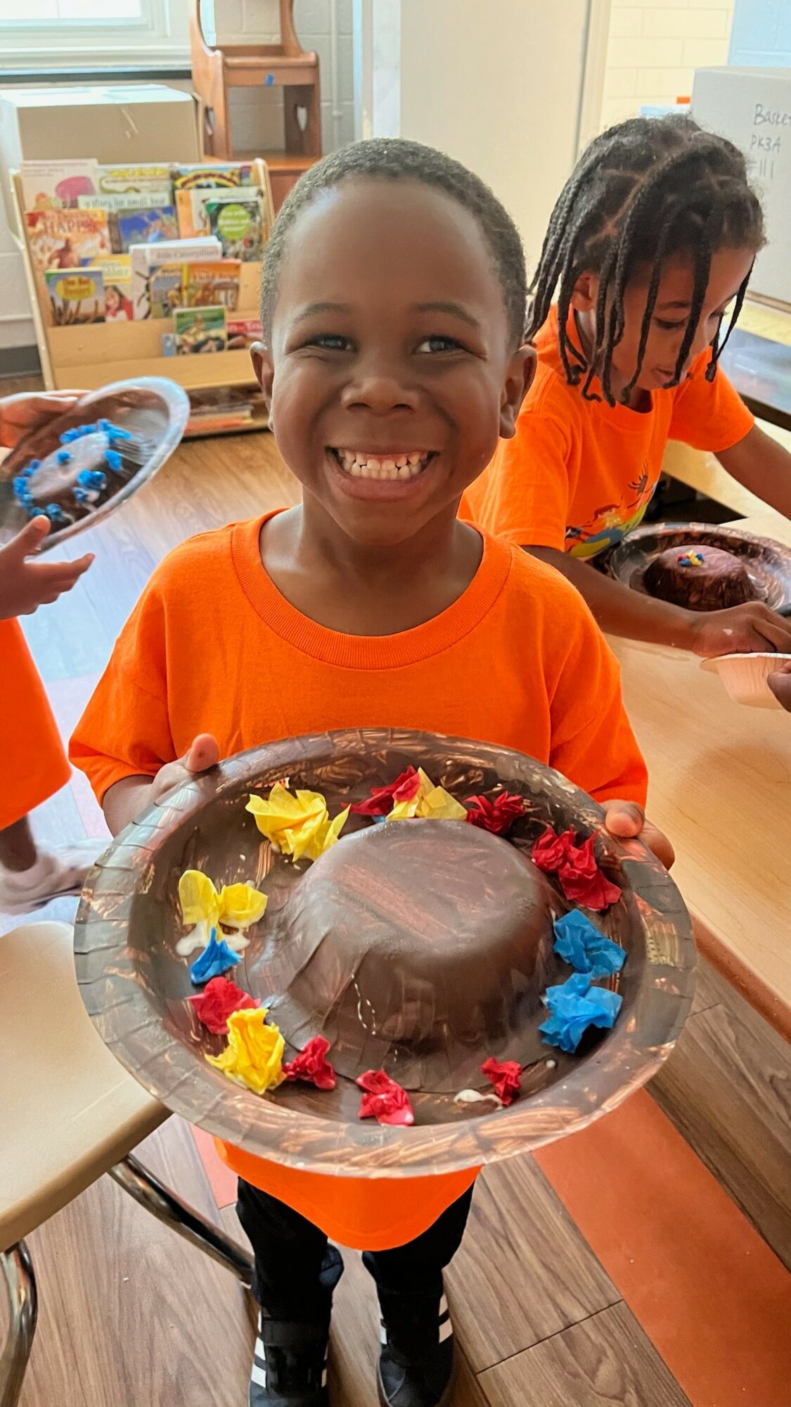 Child smiling and holding a plate with a chocolate cake decorated with colorful flowers, in a classroom setting.