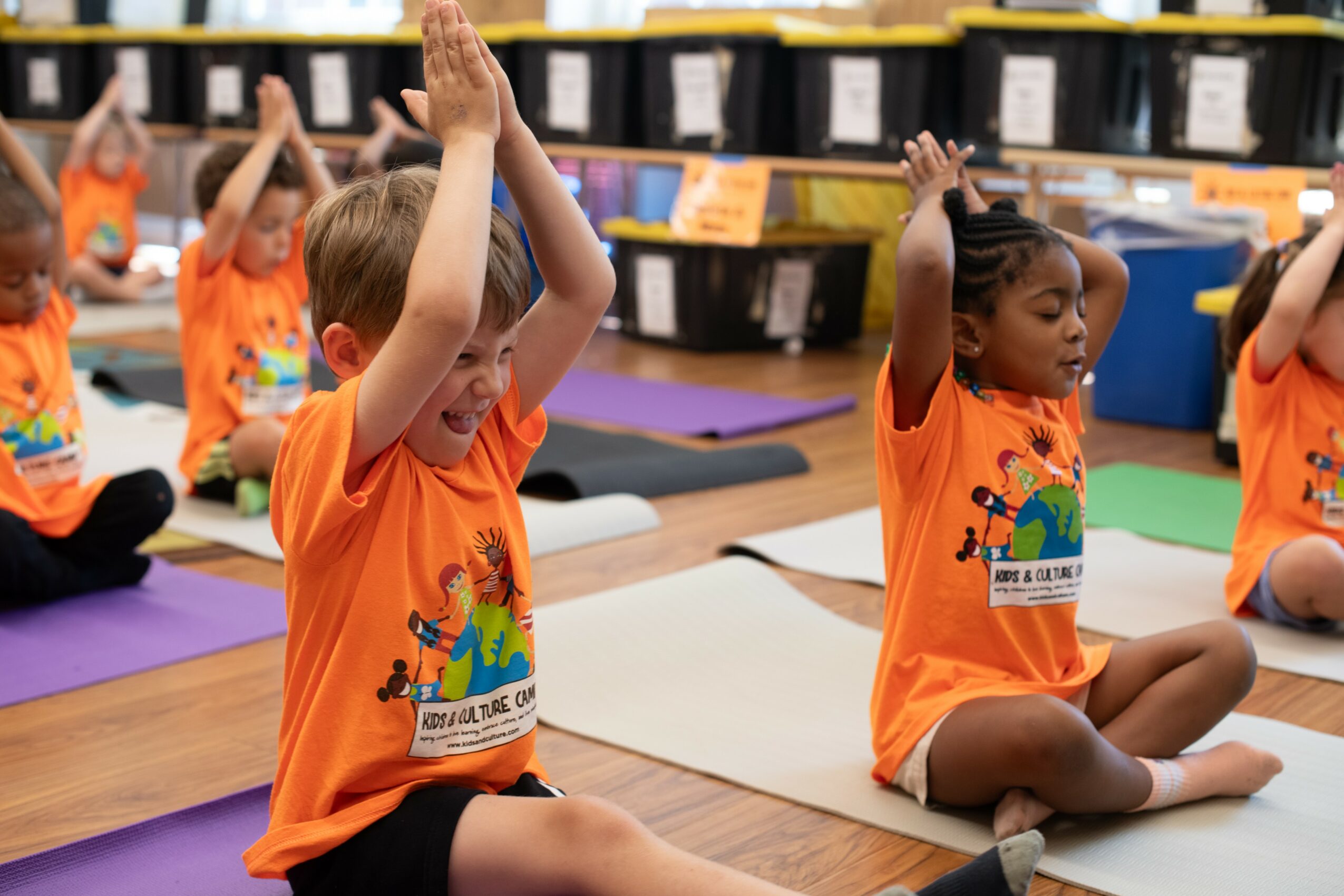 Children sitting on mats in a classroom, participating in a yoga or meditation activity, wearing orange shirts, with bookshelves in the background.
