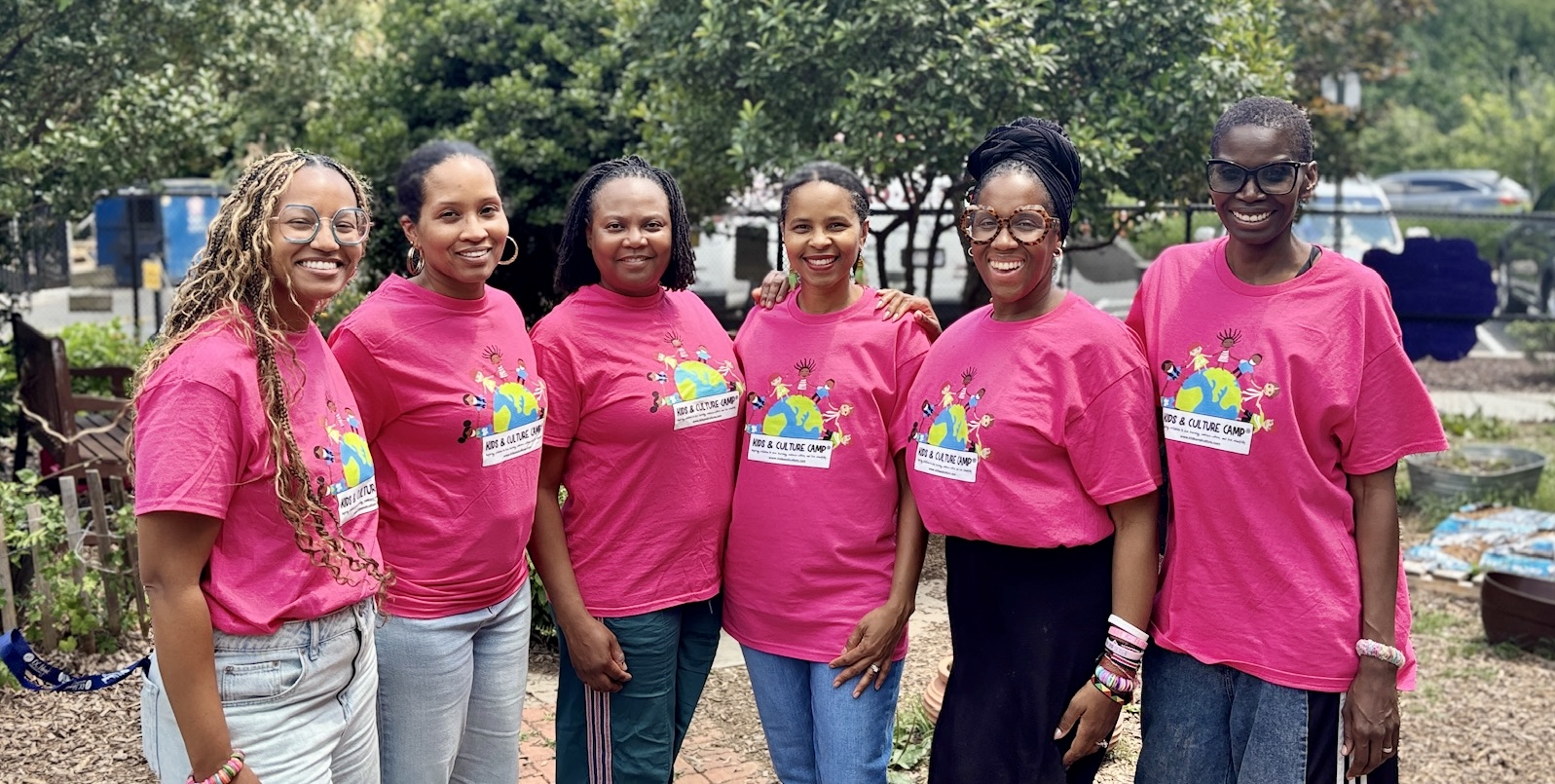 Six women standing outdoors, wearing matching pink T-shirts with a colorful logo, smiling, with trees and cars in the background.