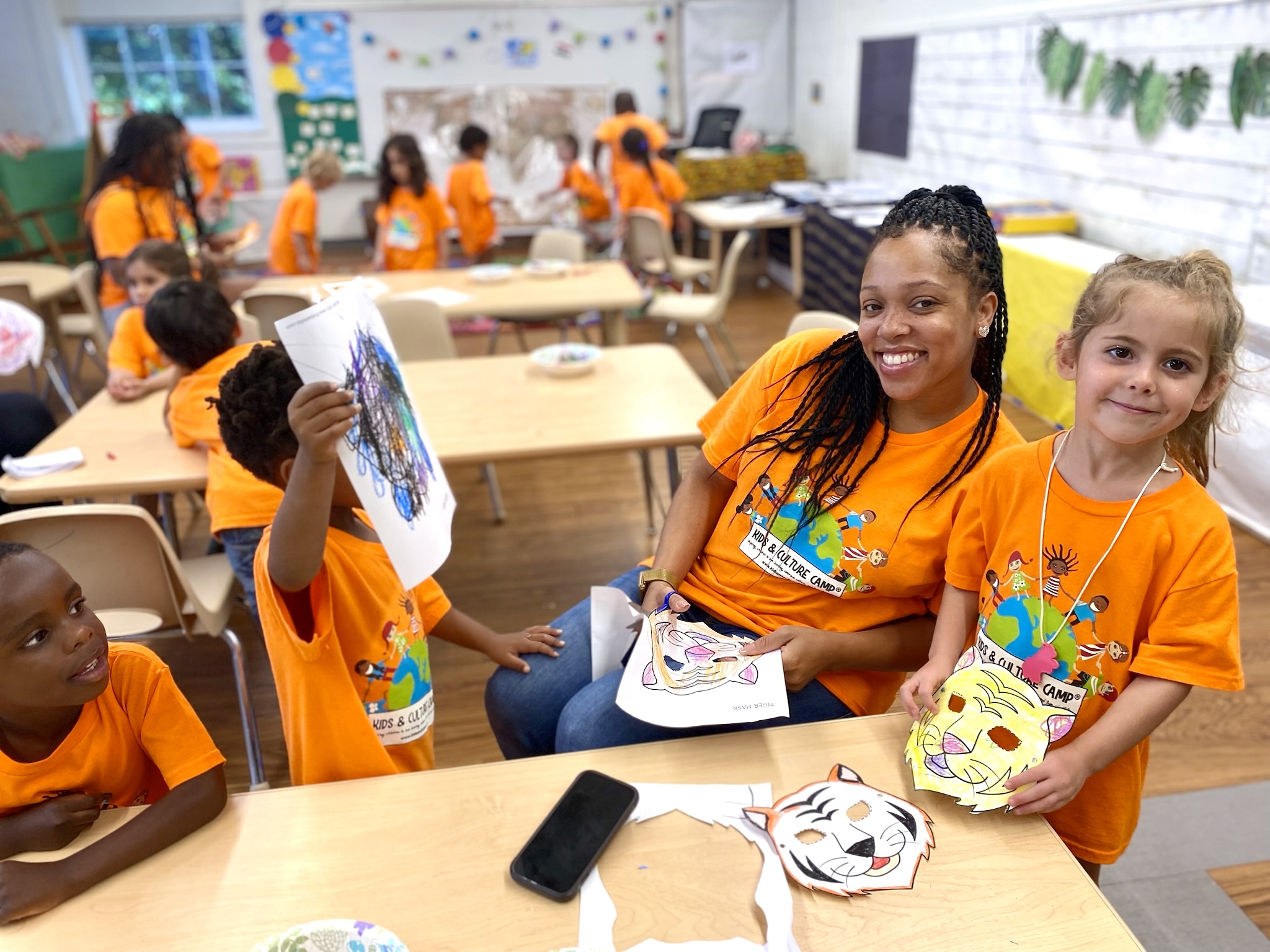 Group of children in orange shirts smiling in classroom, holding colorful paper masks, with desks and educational decorations visible.