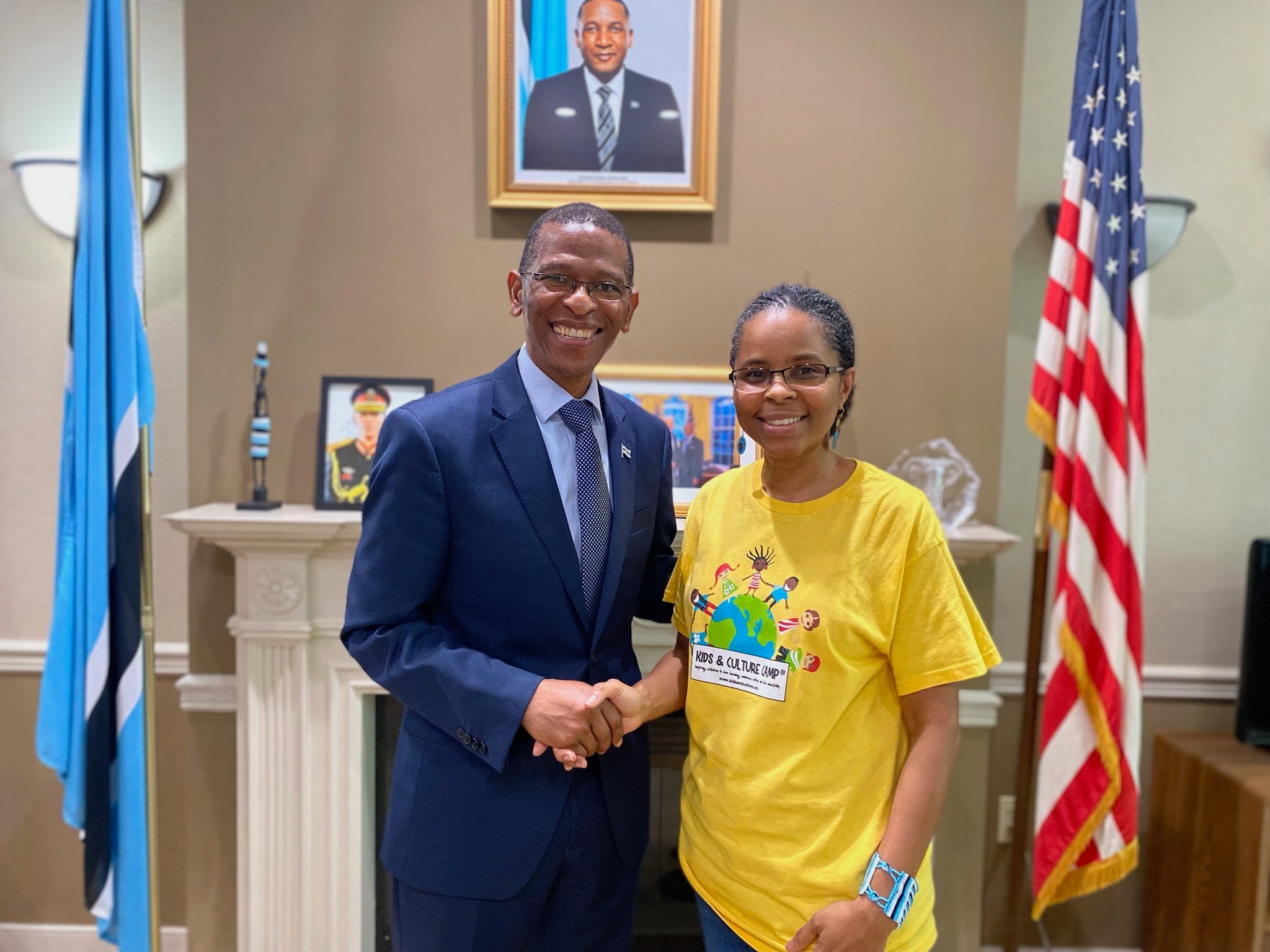 Man in a suit shaking hands with a woman in a yellow T-shirt, both smiling, in a room with flags and a framed photo.