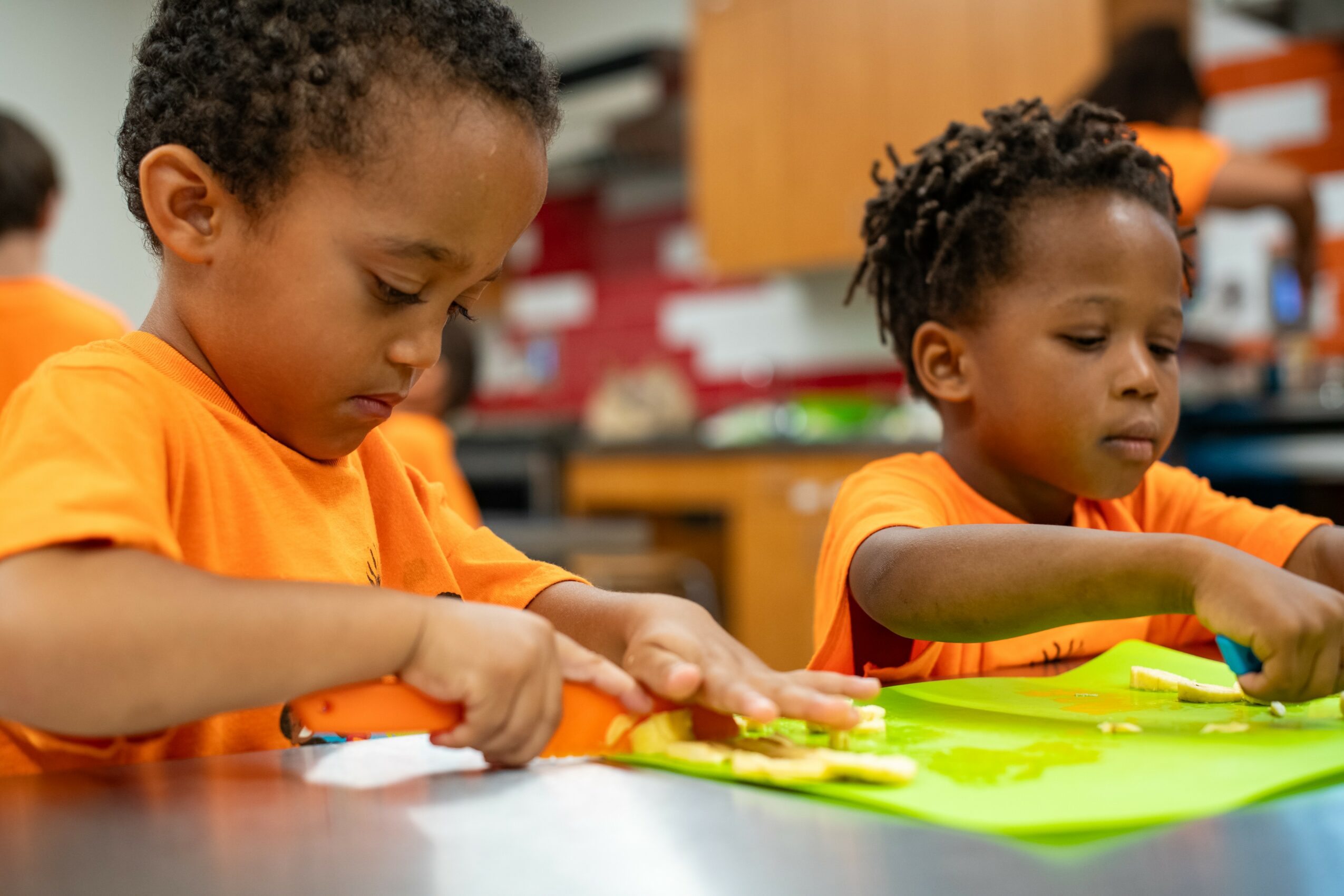 Two children in orange shirts working on a craft project at a table, focused on their tasks.