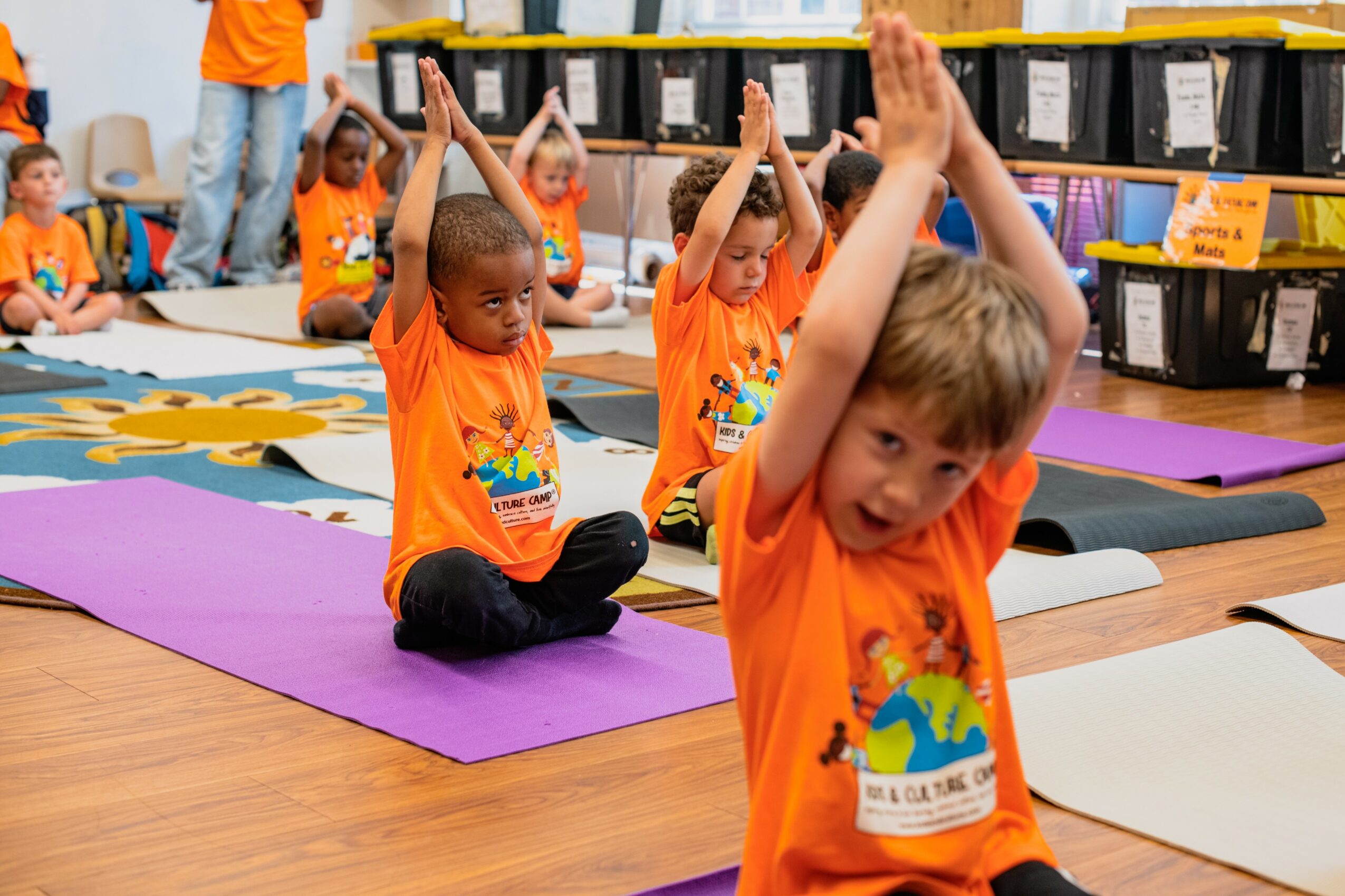 Children in orange shirts sitting on mats, raising hands during a group activity in a classroom.