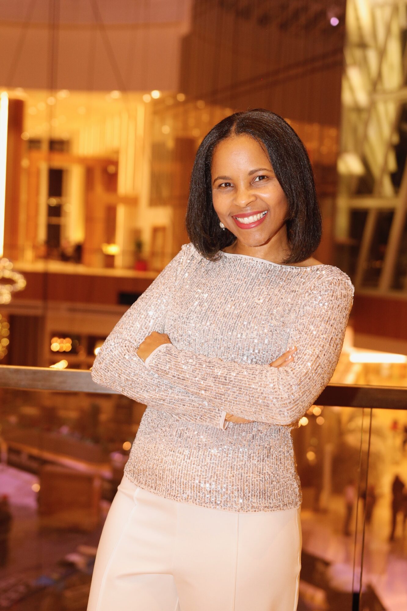 Smiling woman with shoulder-length dark hair, wearing a sparkly long-sleeve top and white pants, standing indoors with arms crossed.