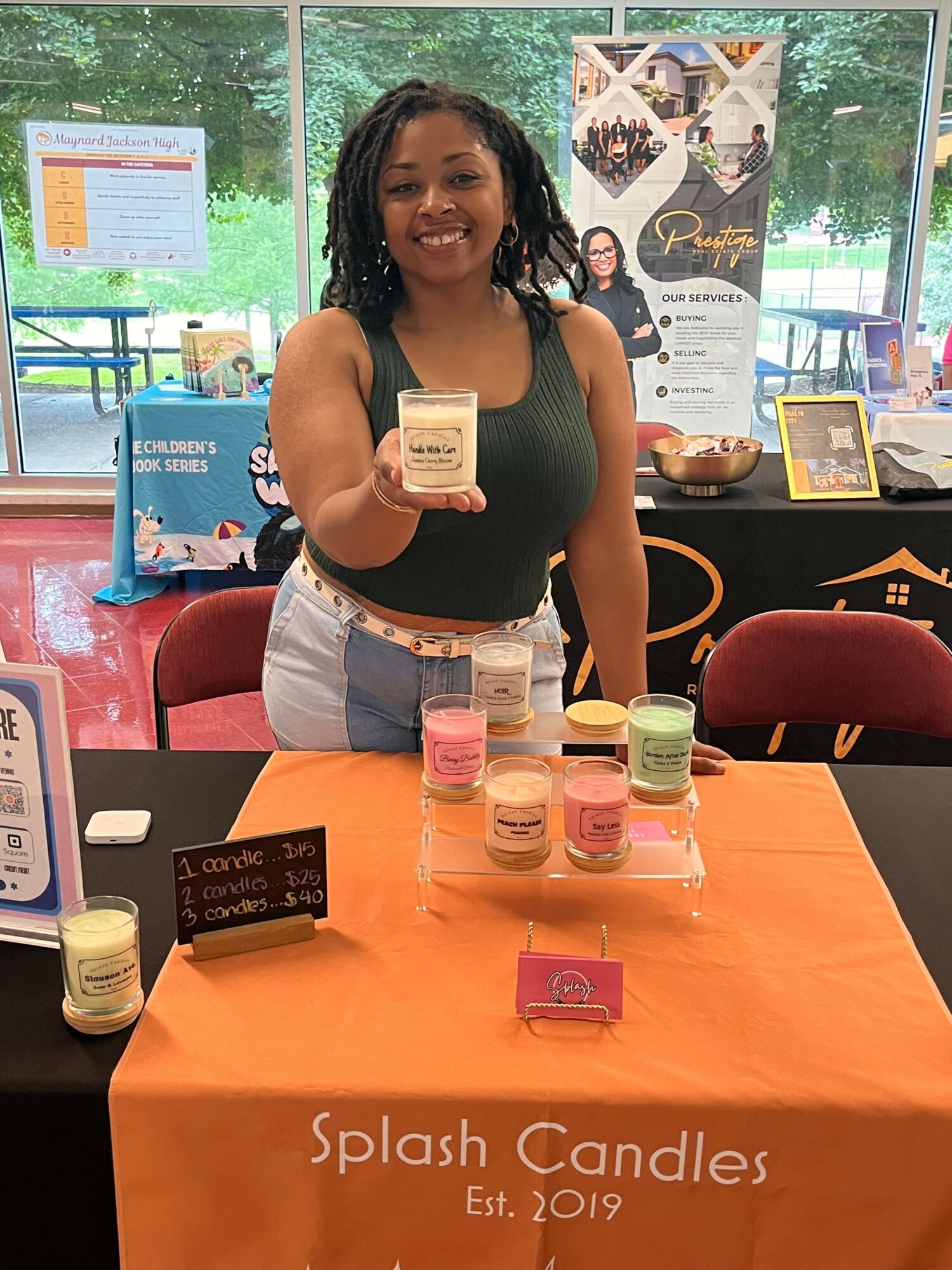 Woman standing behind table with candles and products, indoor setting, window in background, orange tablecloth, promotional display, smiling woman holding a candle, products arranged on table.