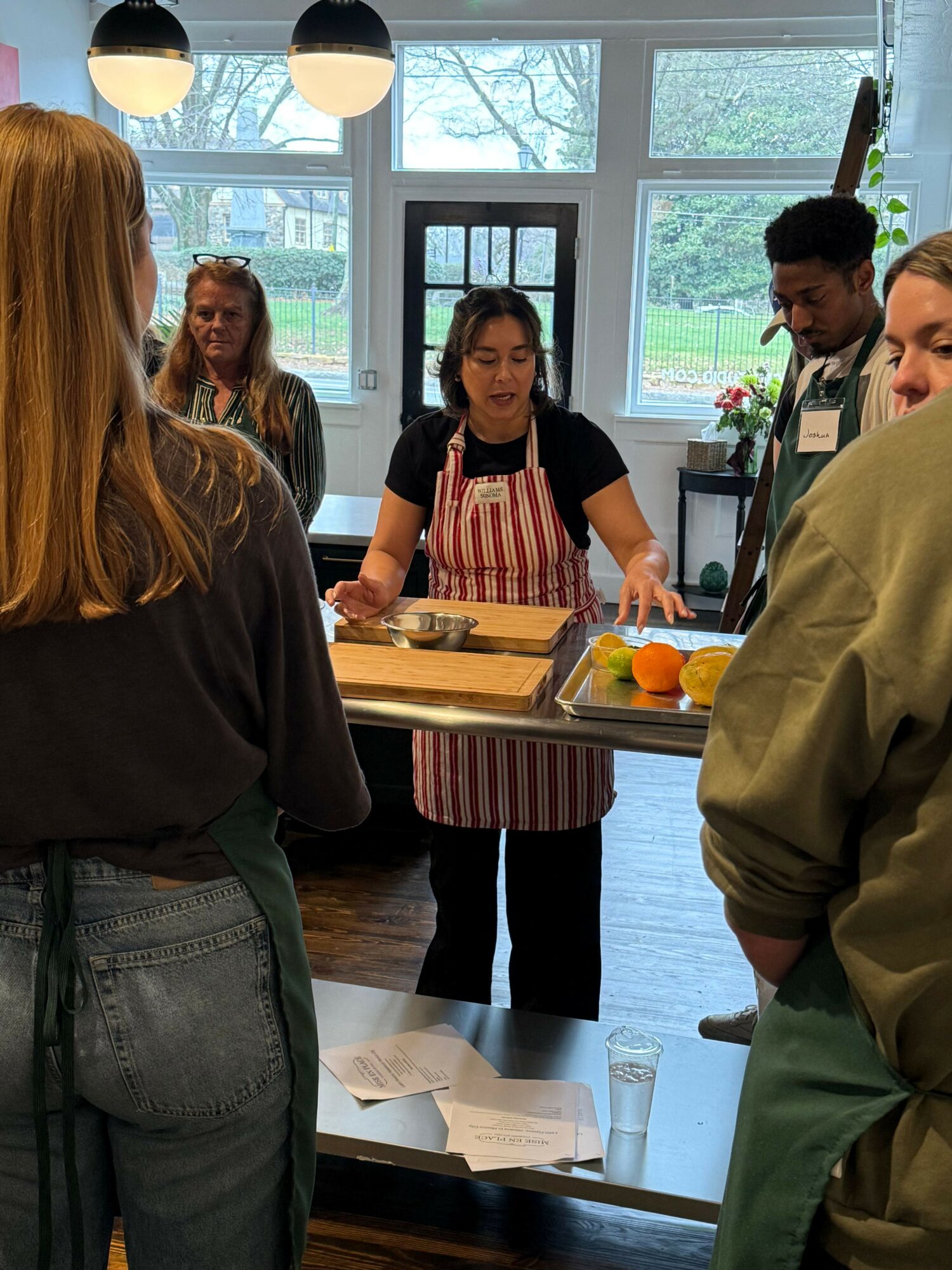 People gathered around a table in a room with large windows, a woman in a striped dress preparing food, others observing.