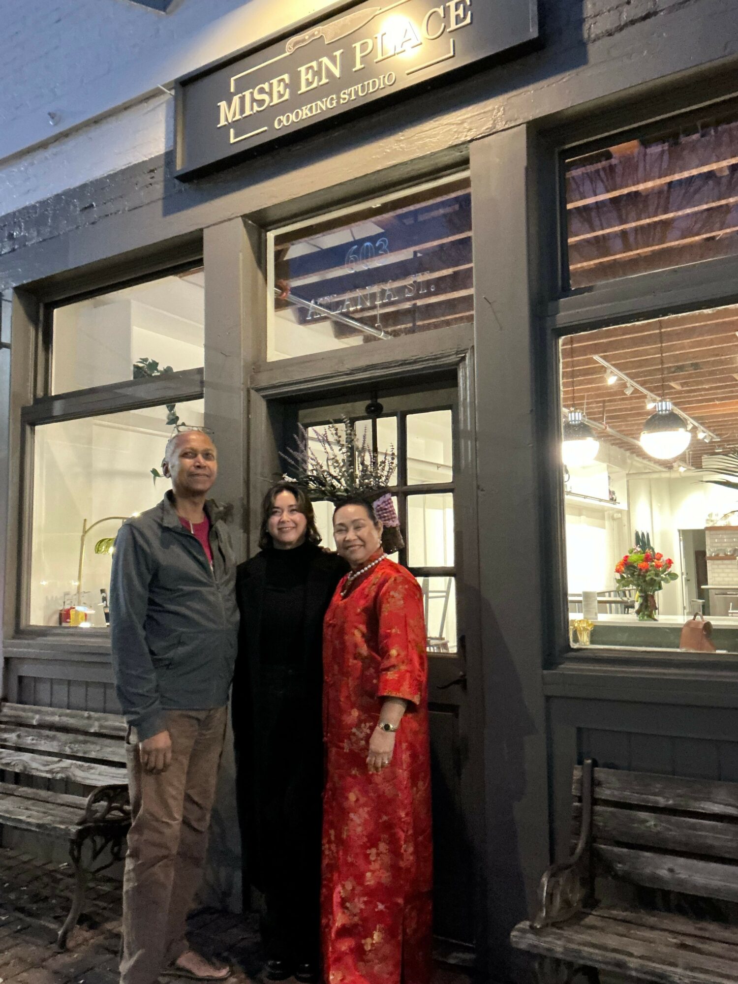 Three people standing outside a building entrance with glass doors, two men and one woman, with a sign above the door.