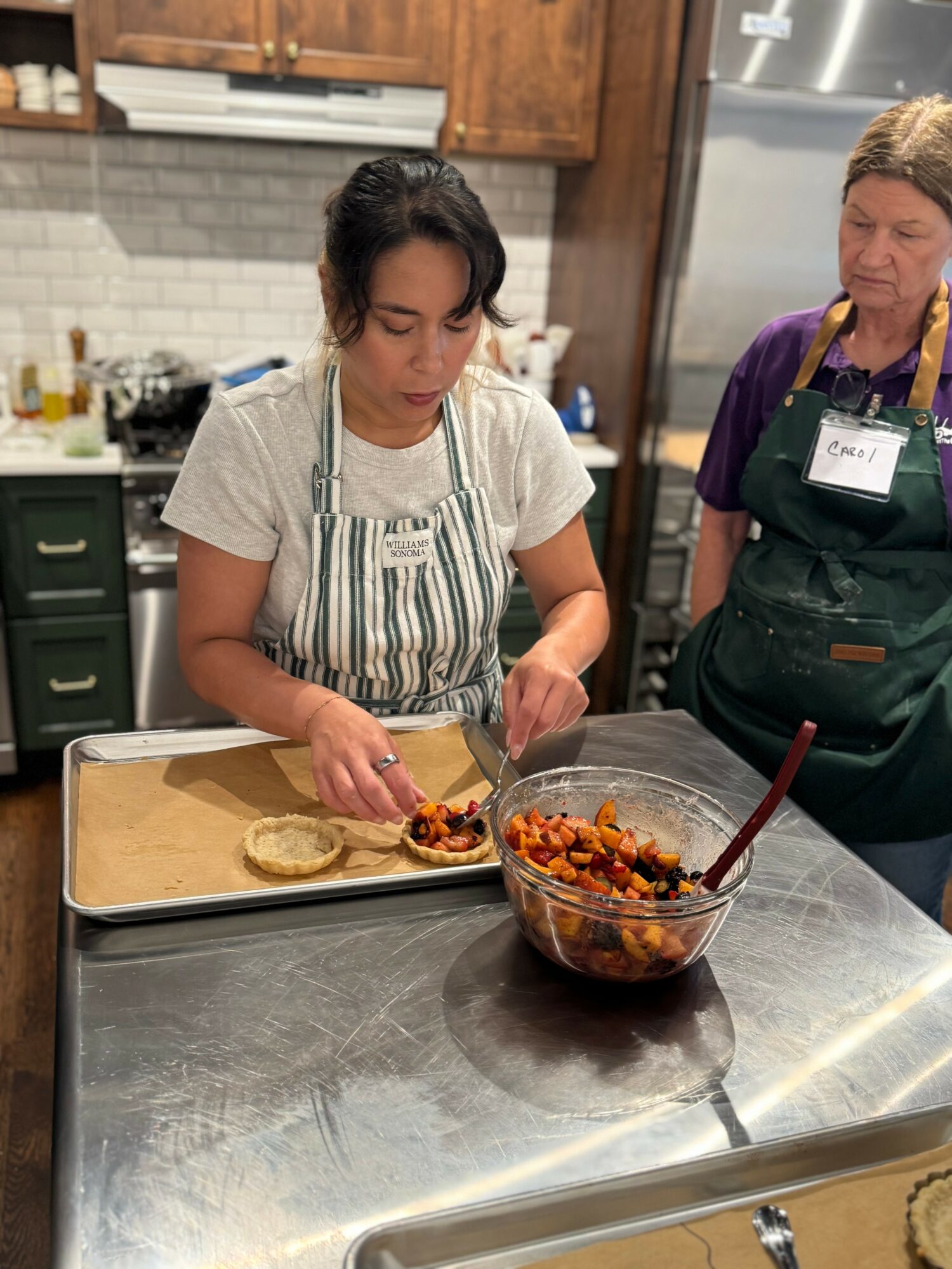Two women preparing food in a kitchen, one filling a tortilla with ingredients, the other observing, with a bowl of filling nearby.