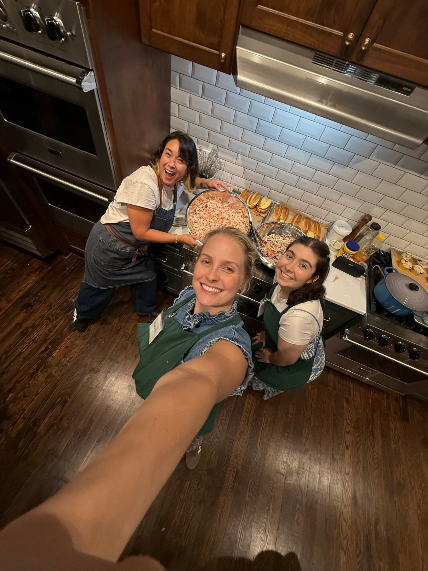 Three people in a kitchen, smiling and taking a selfie, with a stove and white tiled backsplash visible.