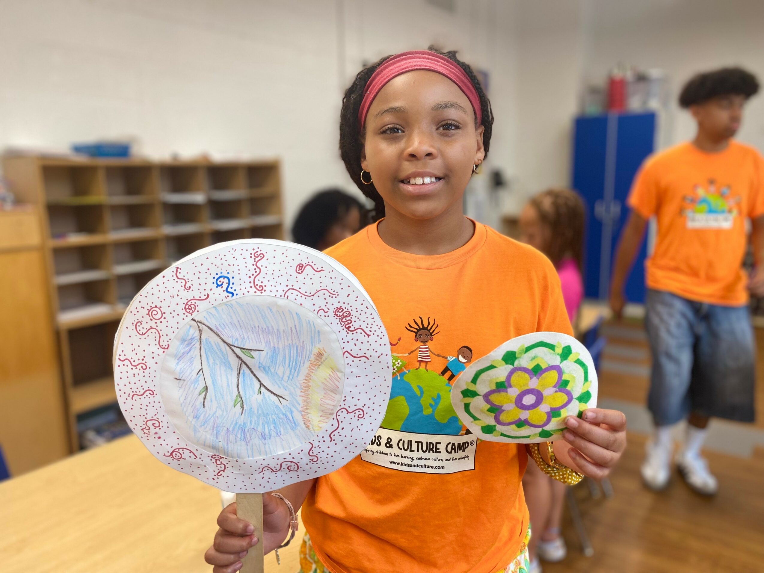 Girl in orange shirt holding decorated paper plate and smaller decorated paper, smiling in classroom with other children in background.