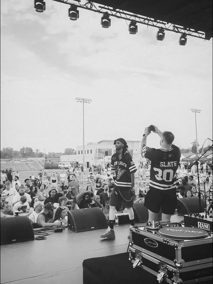 Two performers on stage with a crowd outdoors, one holding a microphone, the other with hands raised, stage equipment visible.