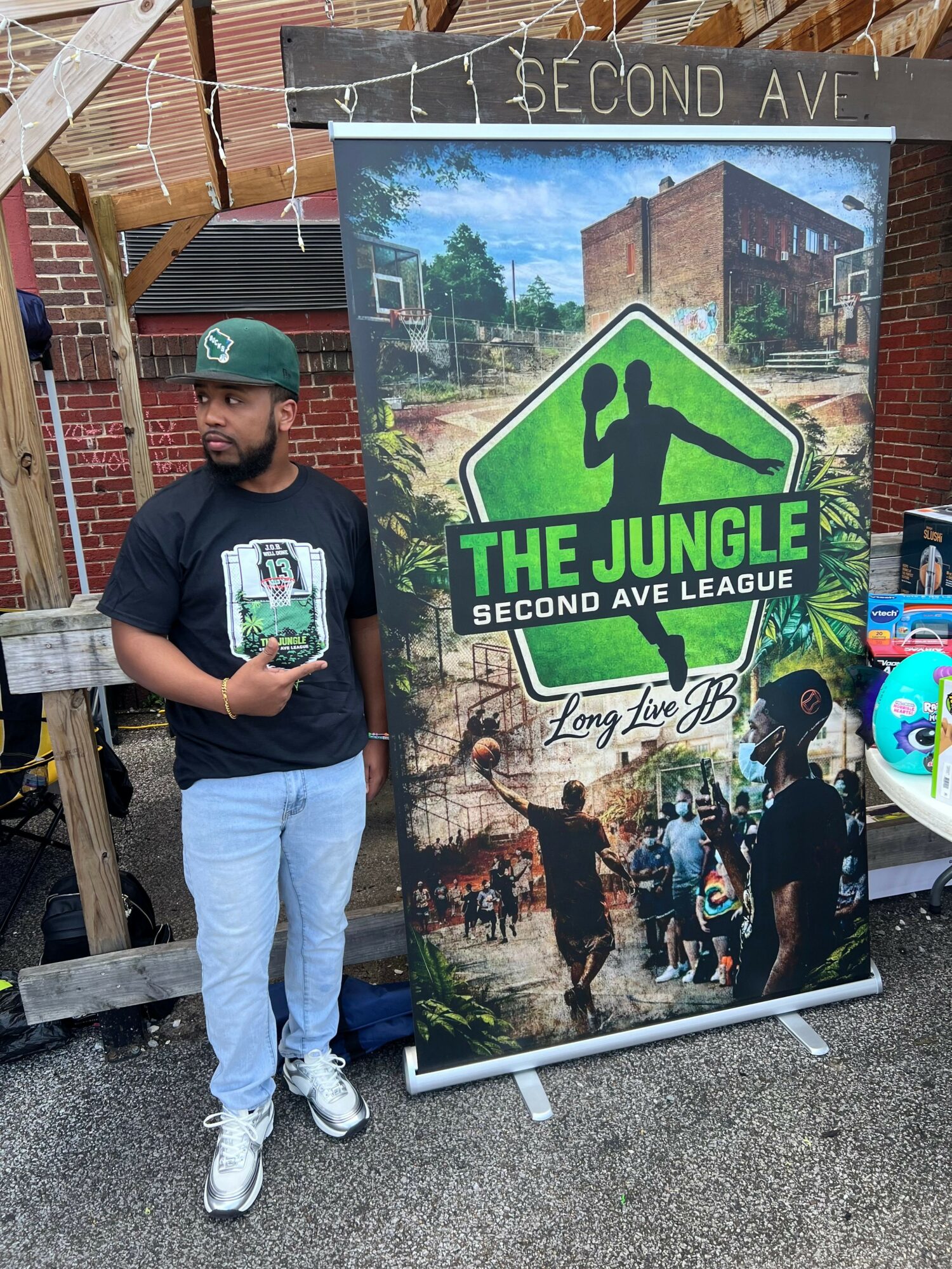 Person standing outdoors holding a brochure in front of a sign for The Jungle Second Day League, with a brick wall and wooden bench nearby.