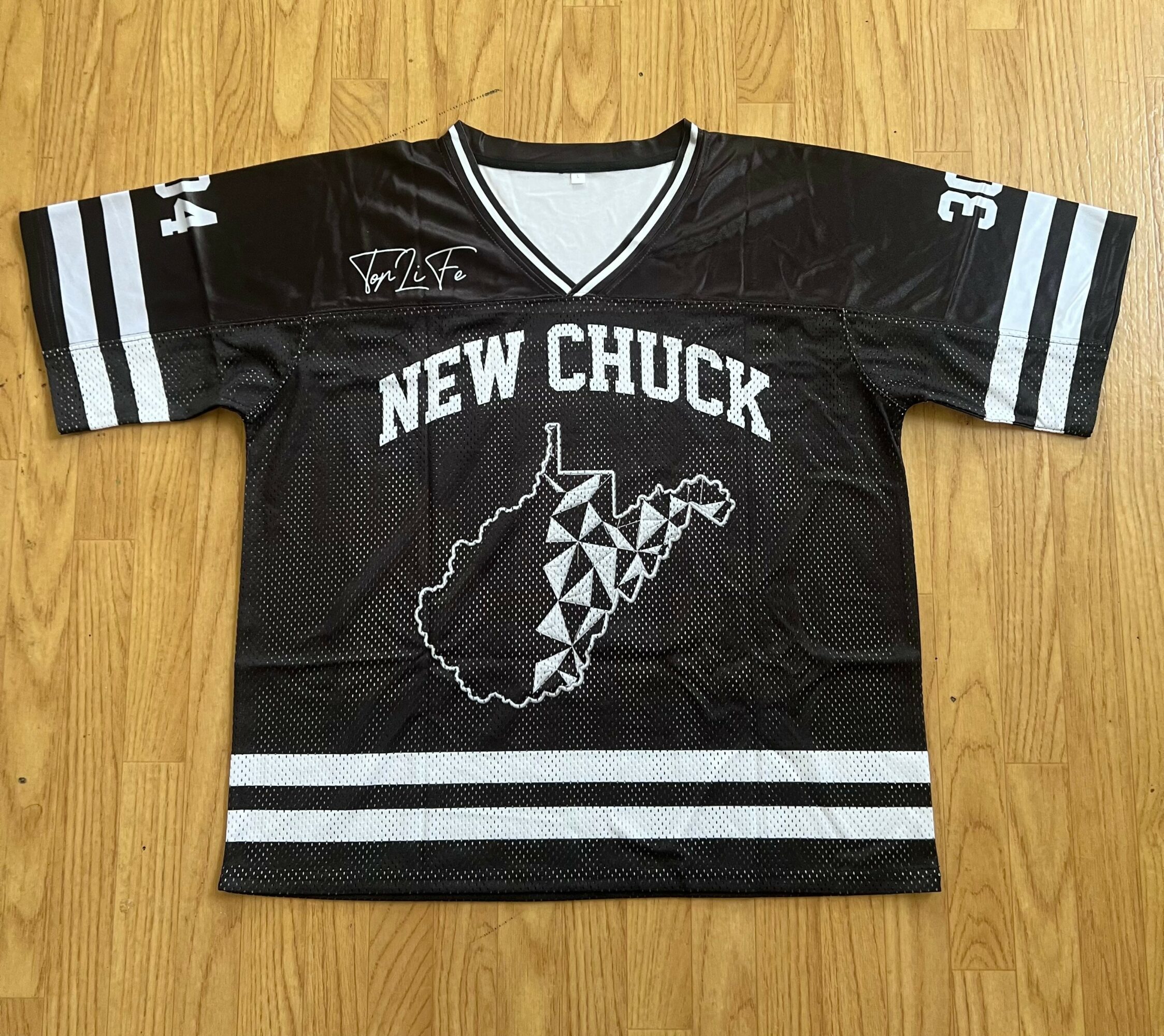 Black sports jersey with white stripes, text 'NEW CHUCK', and a map outline with a geometric pattern, on wooden floor.